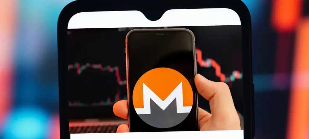 Hand holding a phone displaying the Monero logo over a red candlestick price chart