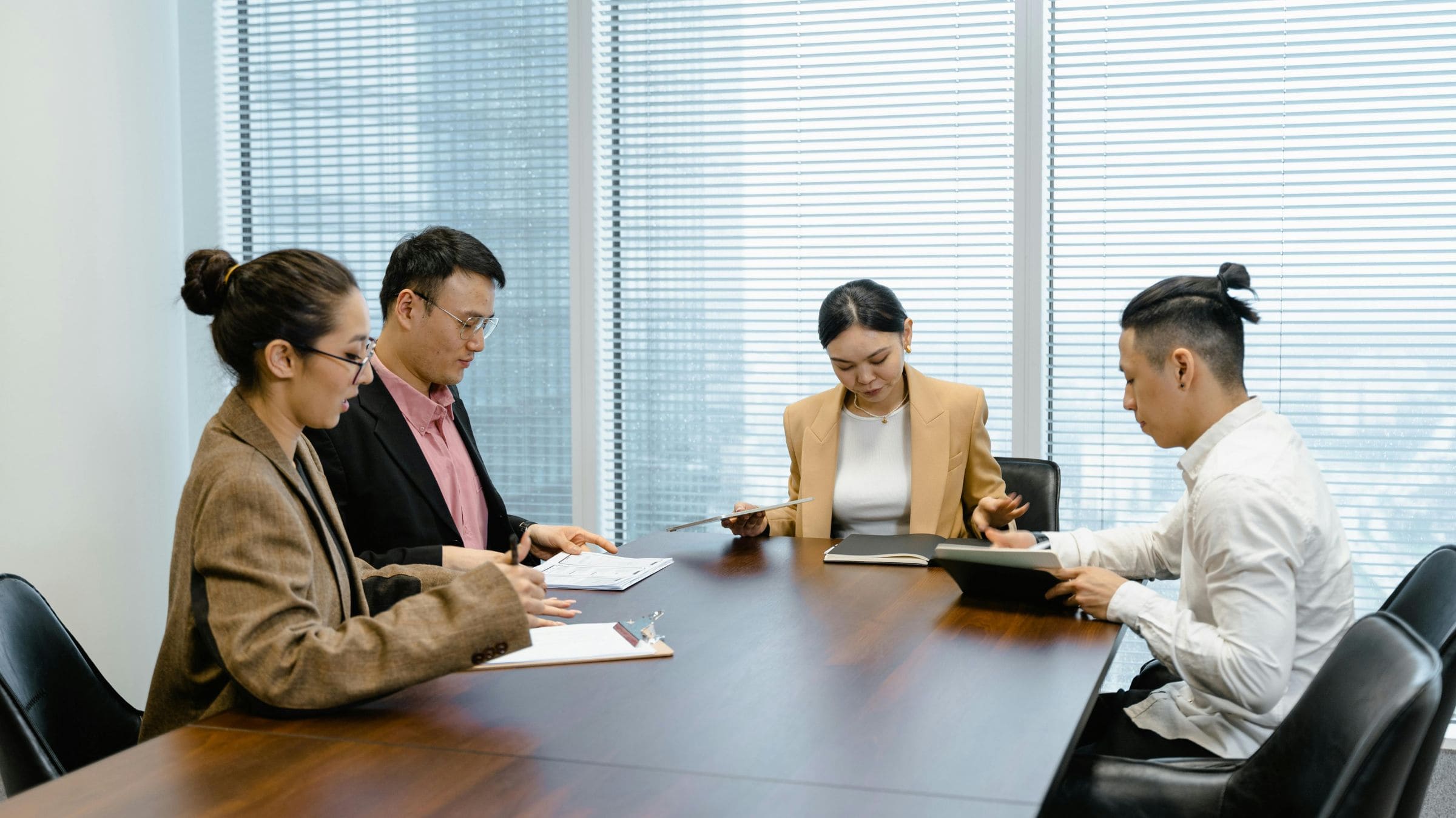 Four office workers sit around a conference table reviewing documents and a tablet in a meeting room.