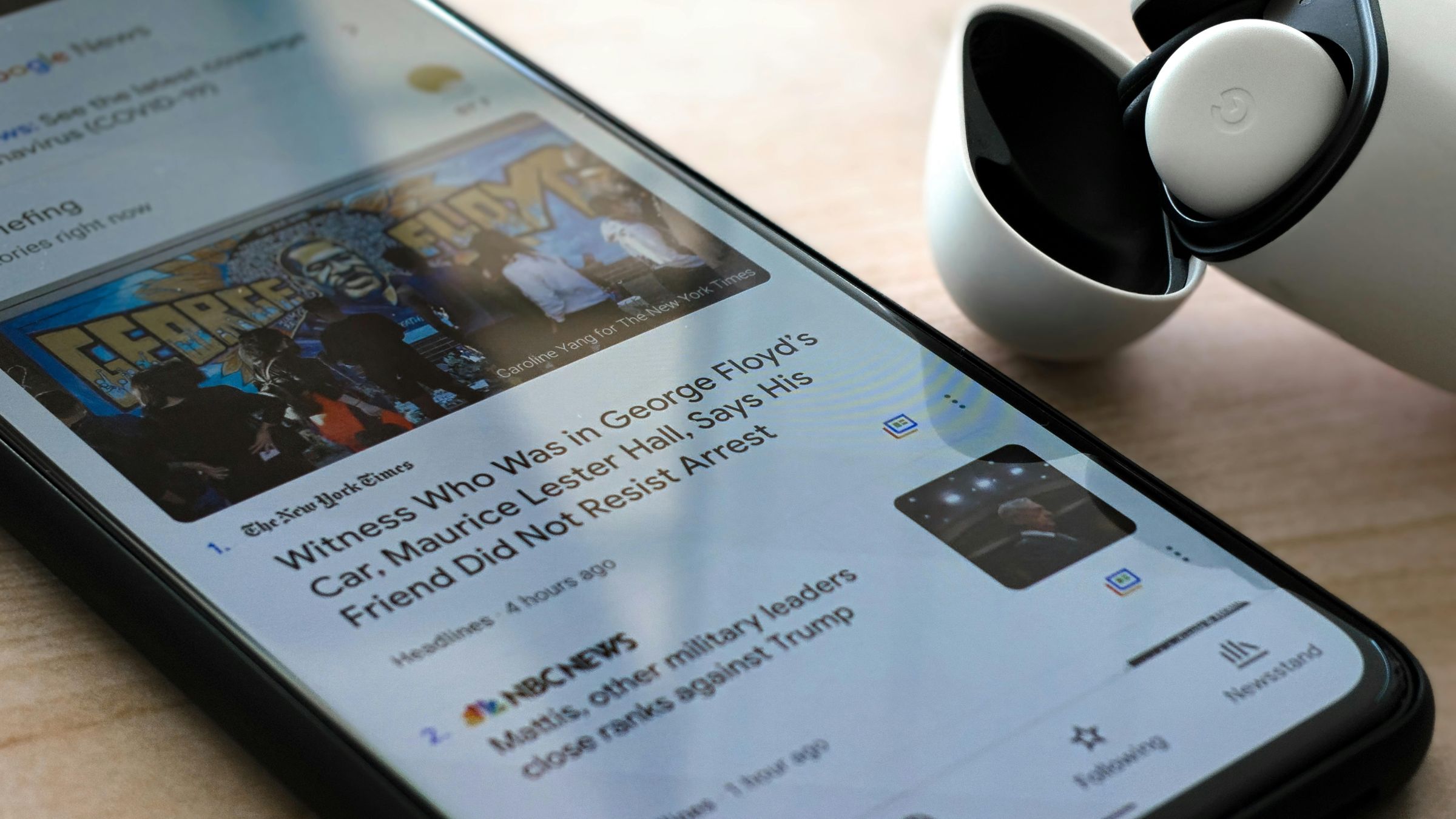 Smartphone displaying Google News headlines beside a wireless earbud case on a tabletop.