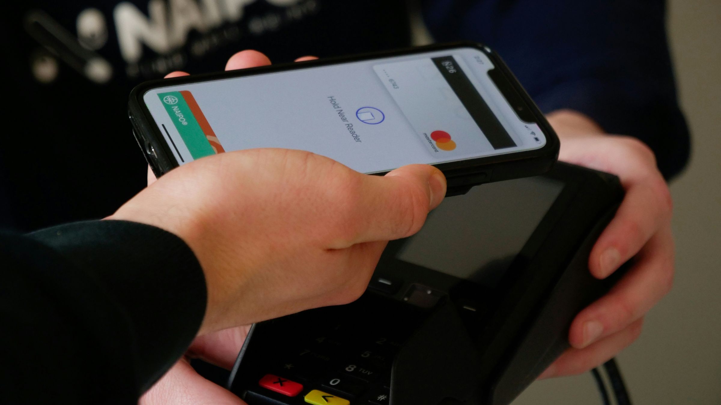 Hand holds a smartphone over a card reader while making a contactless payment at checkout.