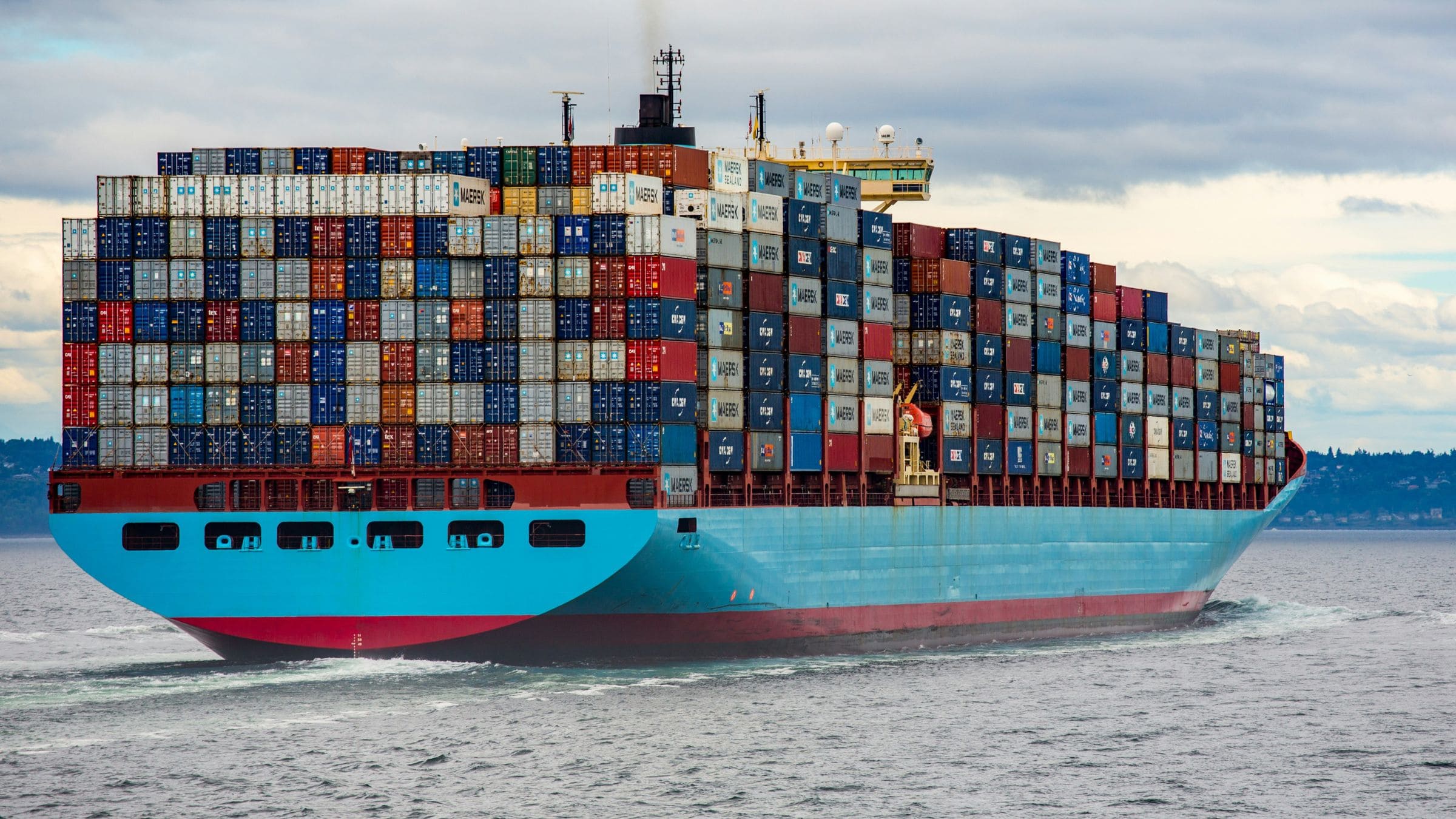 A large container ship carrying stacked cargo containers across open water under a cloudy sky.