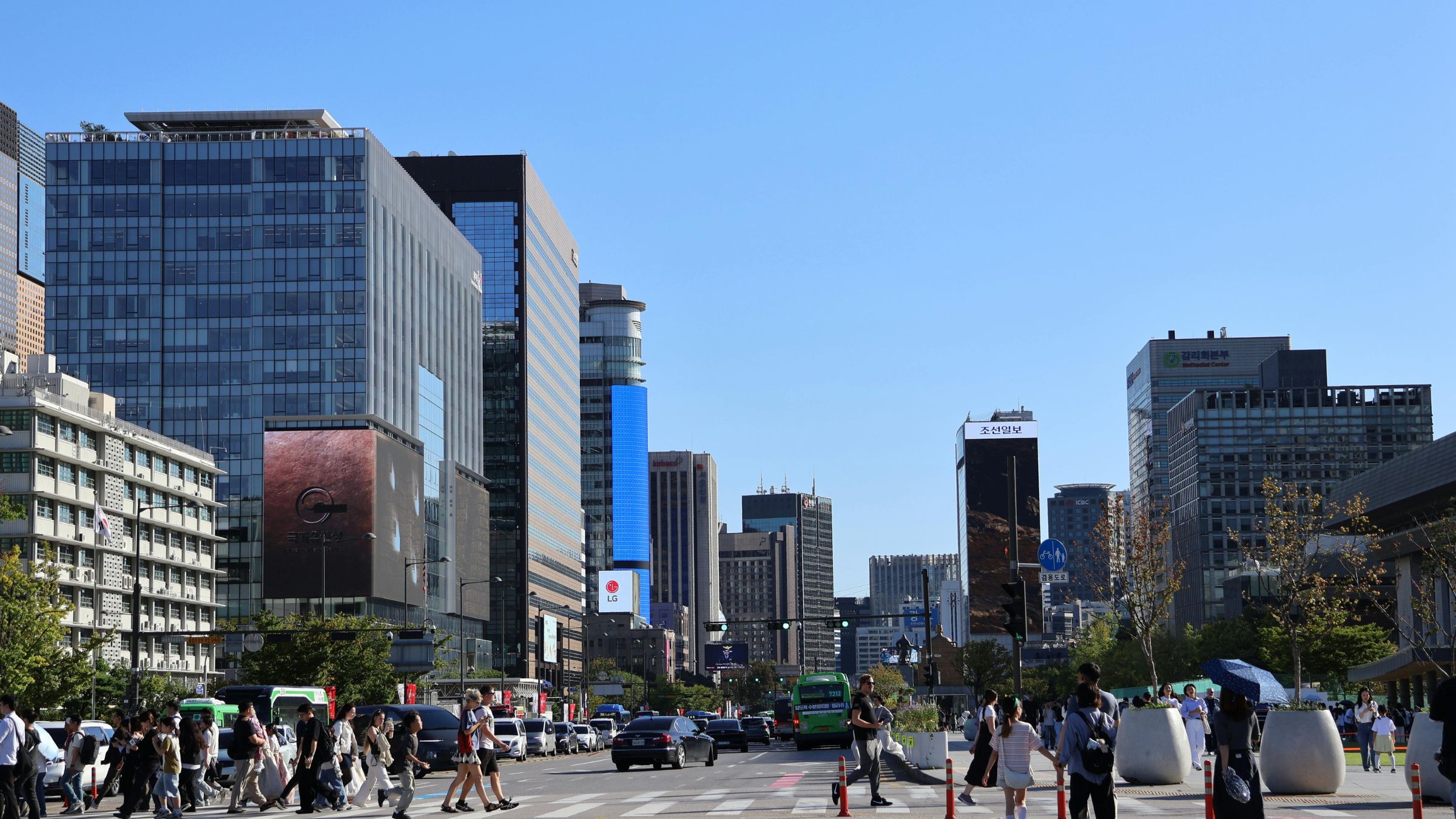 Wide city street with pedestrians, traffic, and modern office buildings under a clear blue sky.