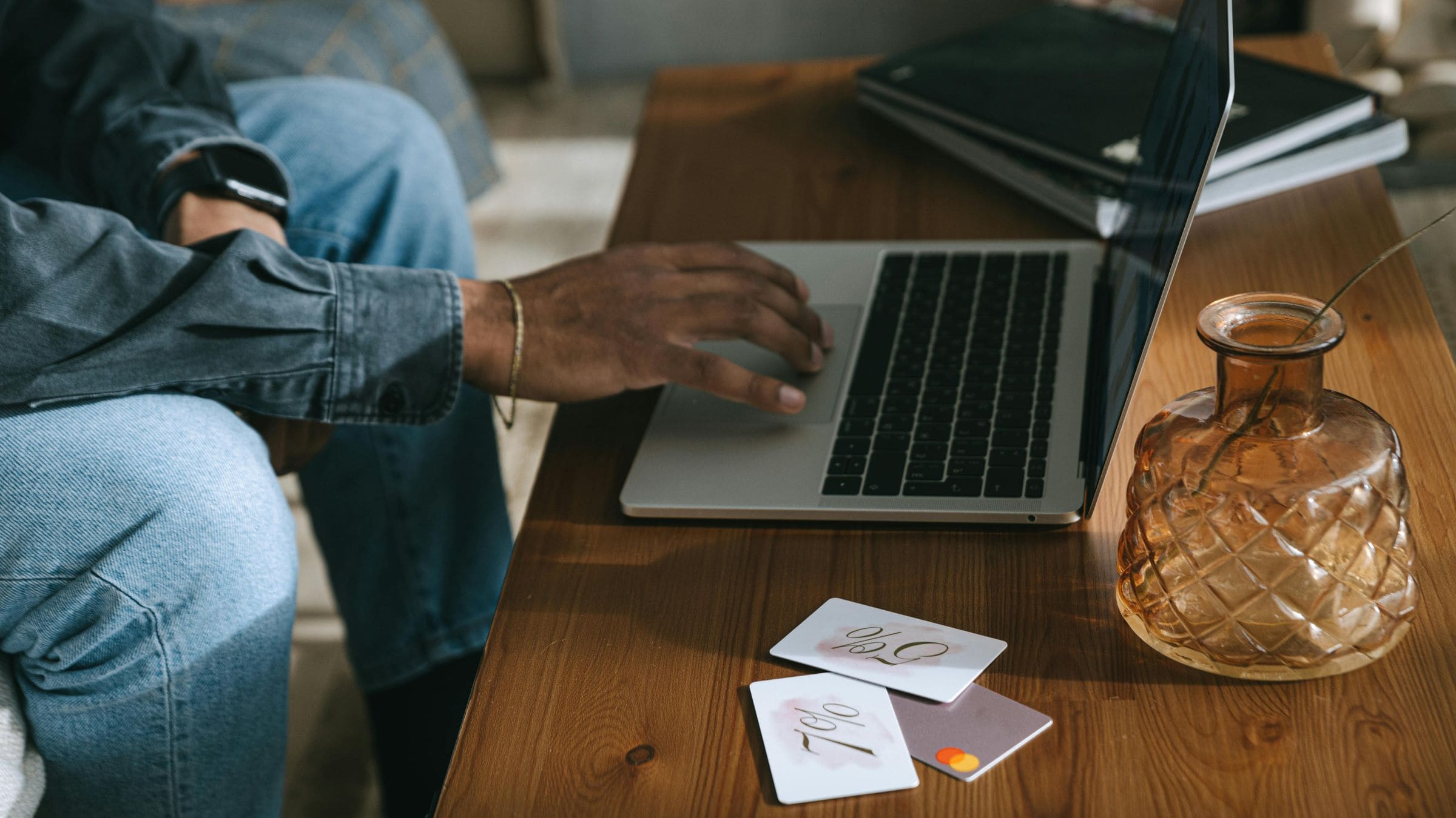 A person using a laptop beside cards and a glass vase on a wooden coffee table.