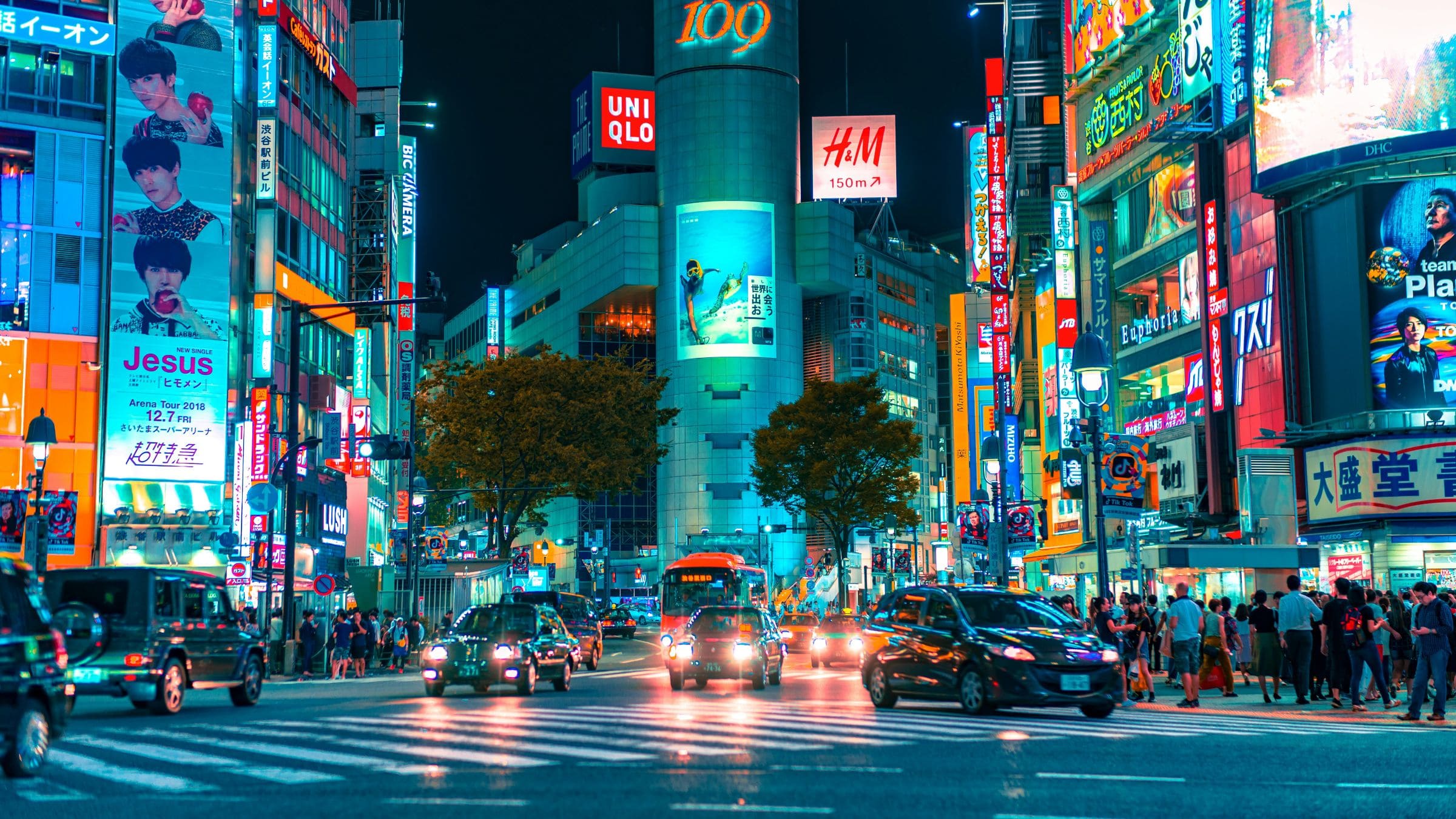 Night view of Shibuya Crossing in Tokyo with traffic, pedestrians, and brightly lit commercial billboards.