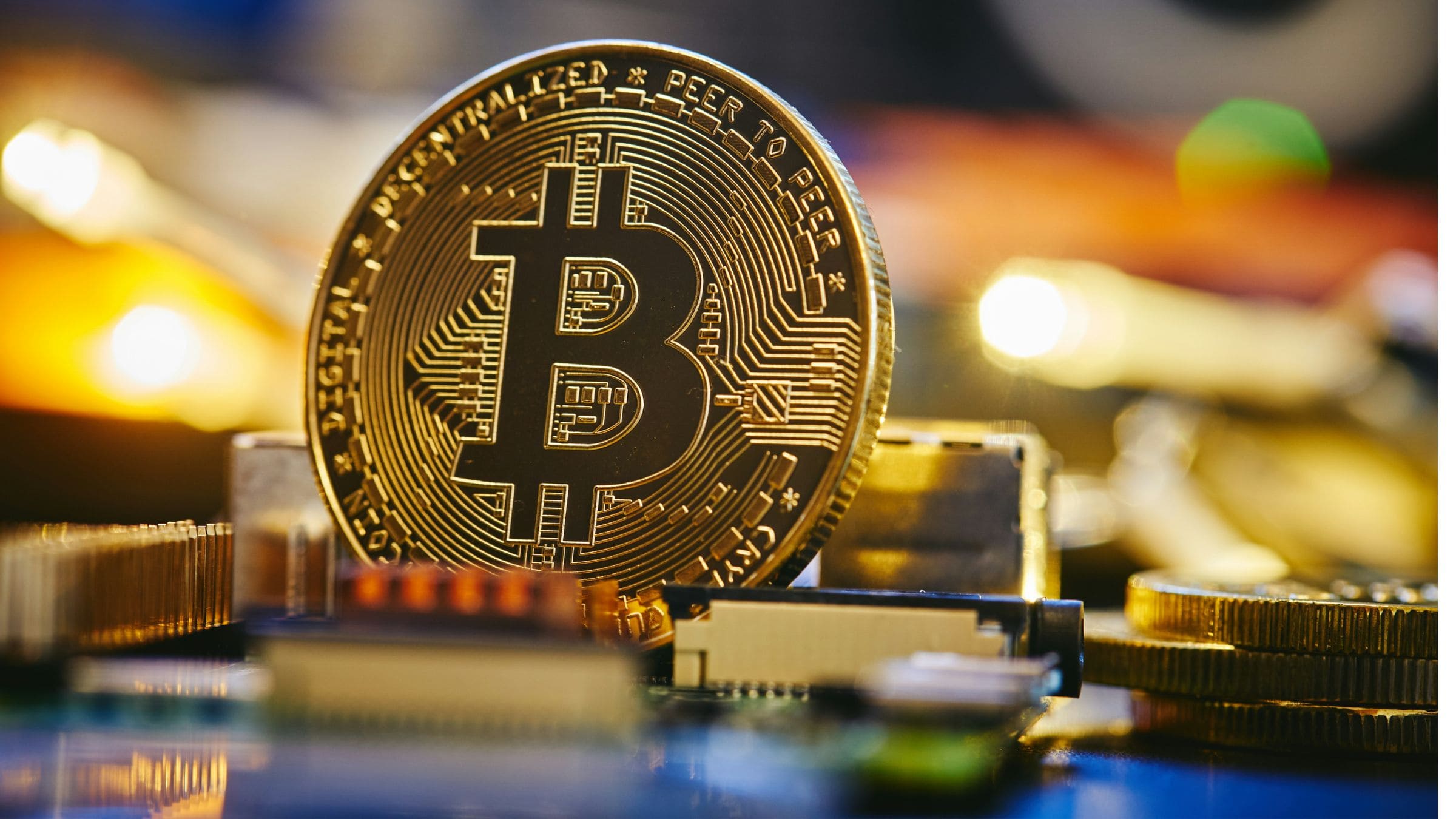 Gold-colored Bitcoin token displayed on computer hardware and electronic components with blurred lights in background.