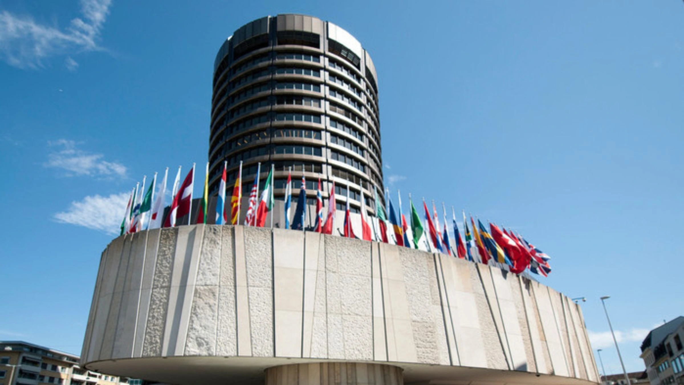 The cylindrical tower of the Bank for International Settlements headquarters in Basel, Switzerland, surrounded by flags of member nations against a clear blue sky.