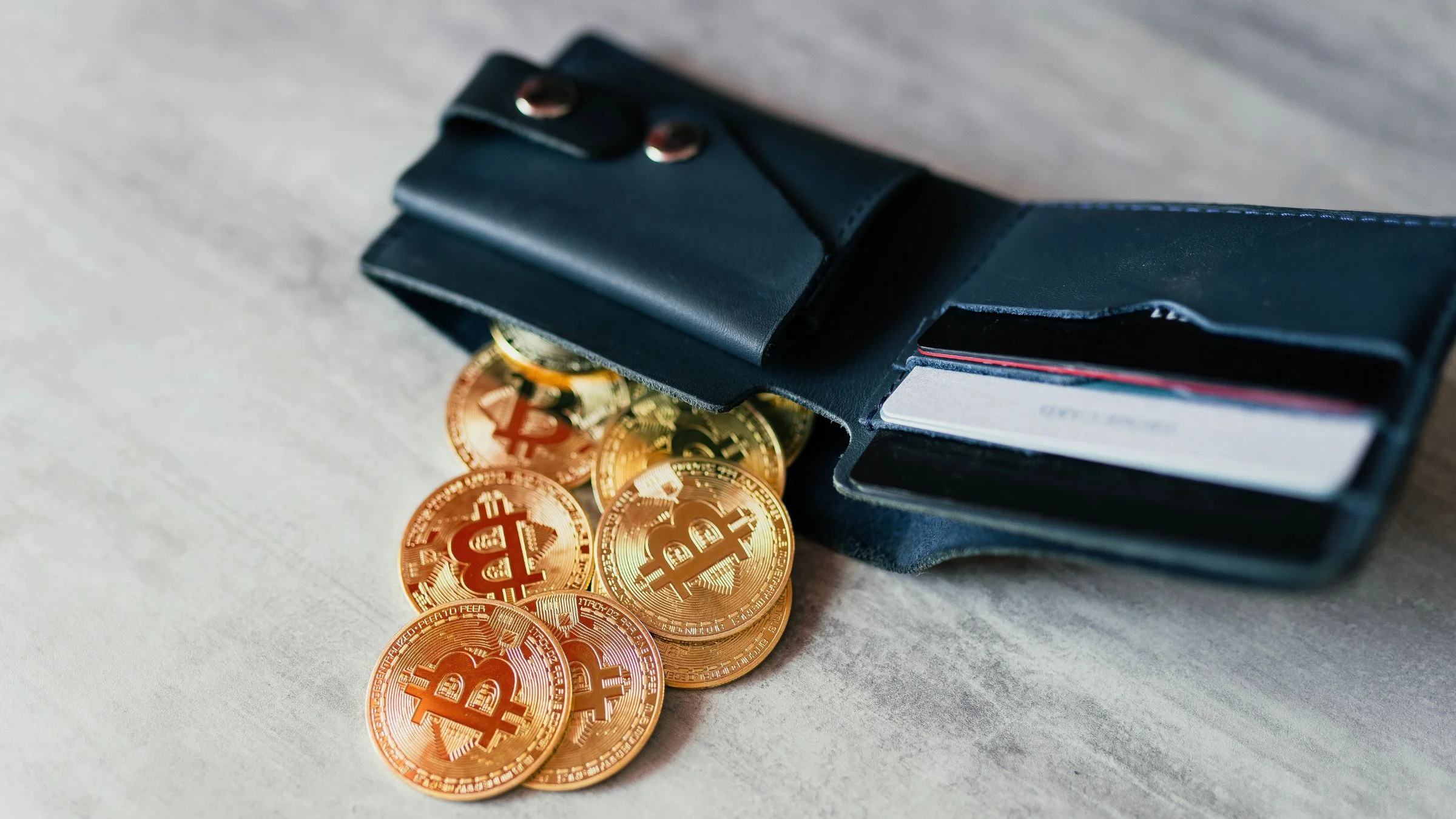 Multiple golden Bitcoin coins spilling out of a dark leather wallet with payment cards on a gray surface.