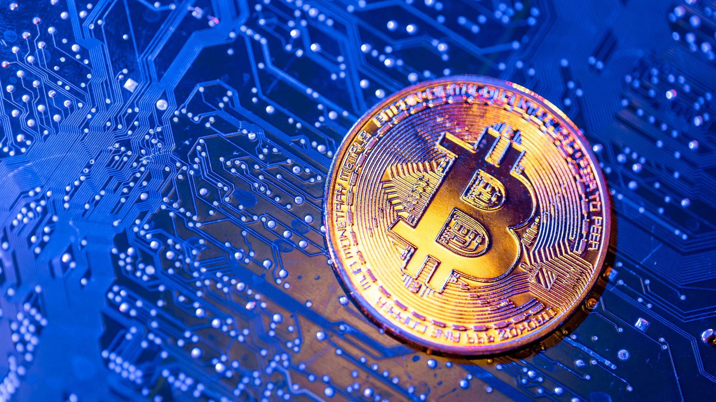 A golden Bitcoin coin resting on a blue illuminated circuit board with visible electronic pathways and solder points.