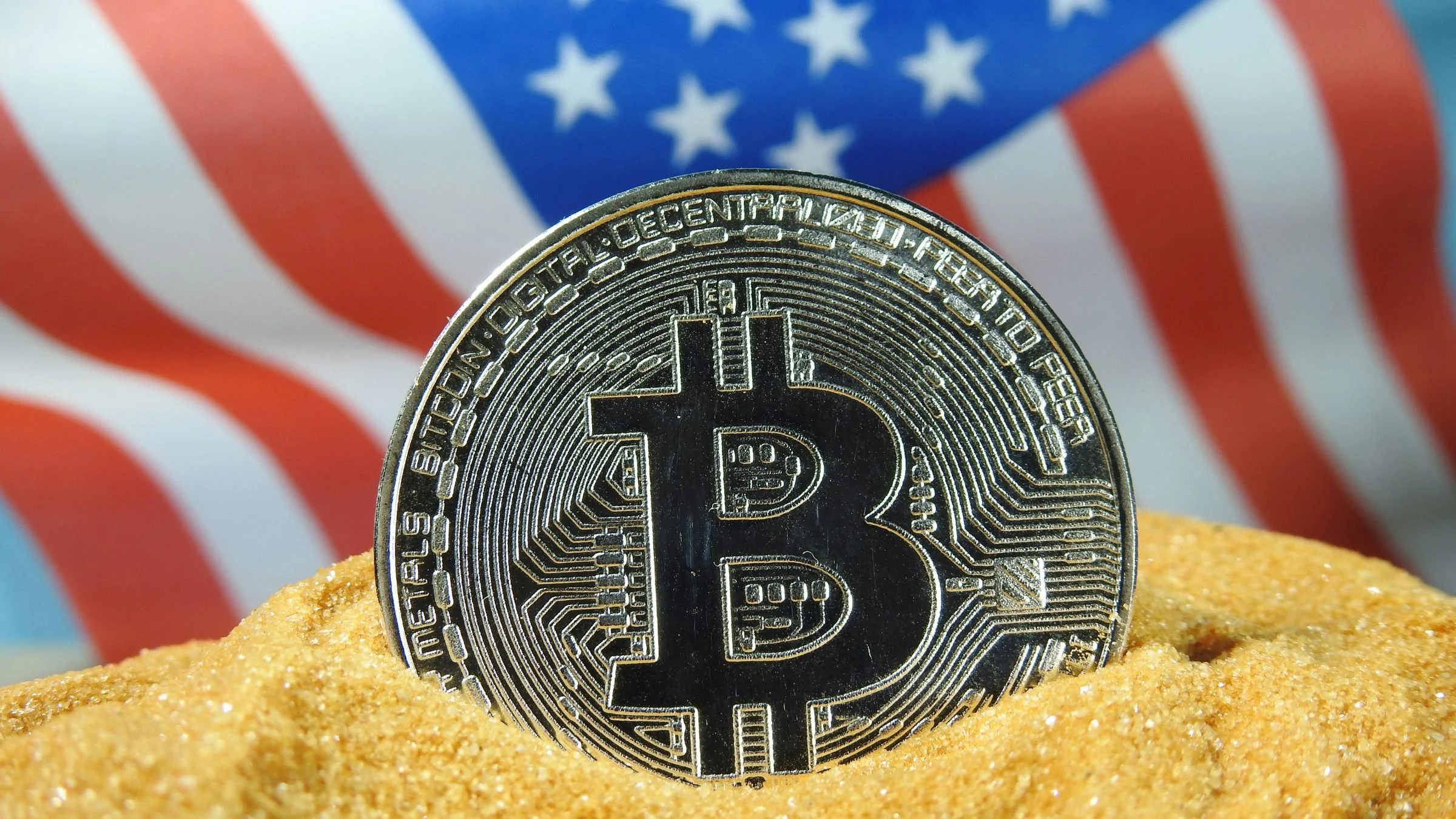 Bitcoin token partly buried in sand with a blurred United States flag in the background.