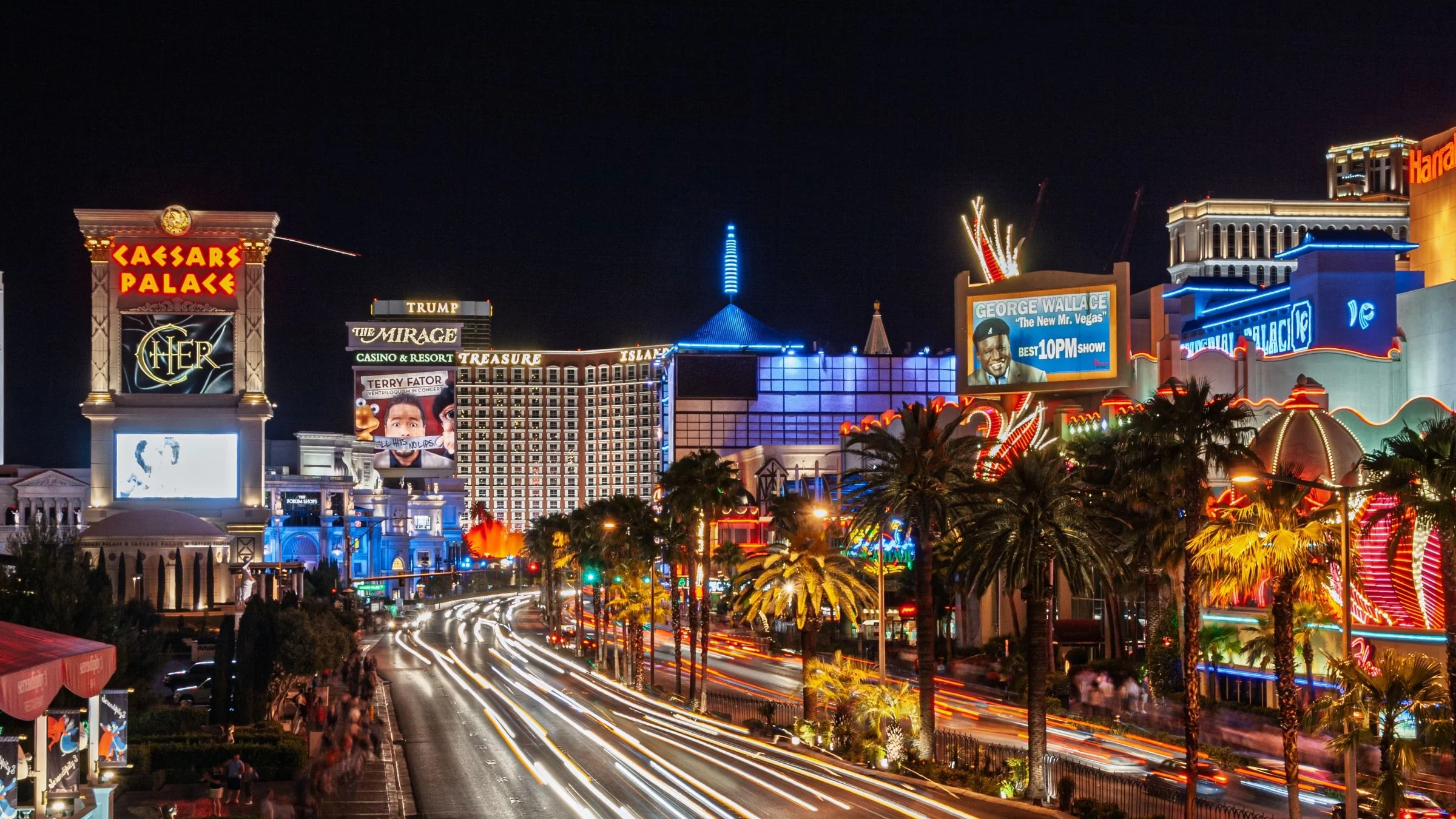 A vibrant nighttime view of the Las Vegas Strip featuring Caesars Palace, The Mirage, and Trump Tower with light trails from passing traffic