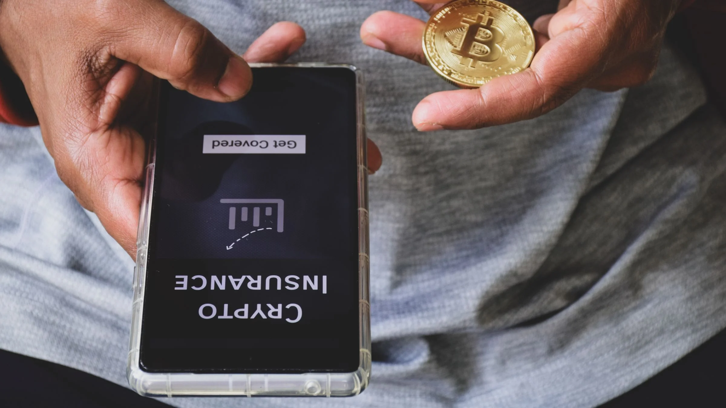Hands holding a Bitcoin coin next to a smartphone displaying "Crypto Insurance" and "Get Covered"