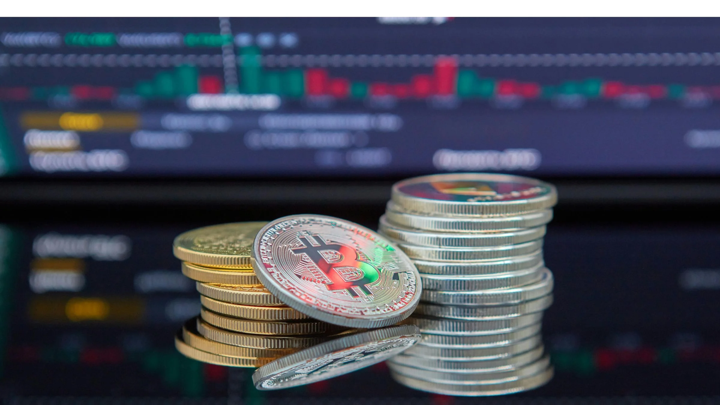 Stack of physical crypto-themed coins in front of a blurred trading screen with market charts.