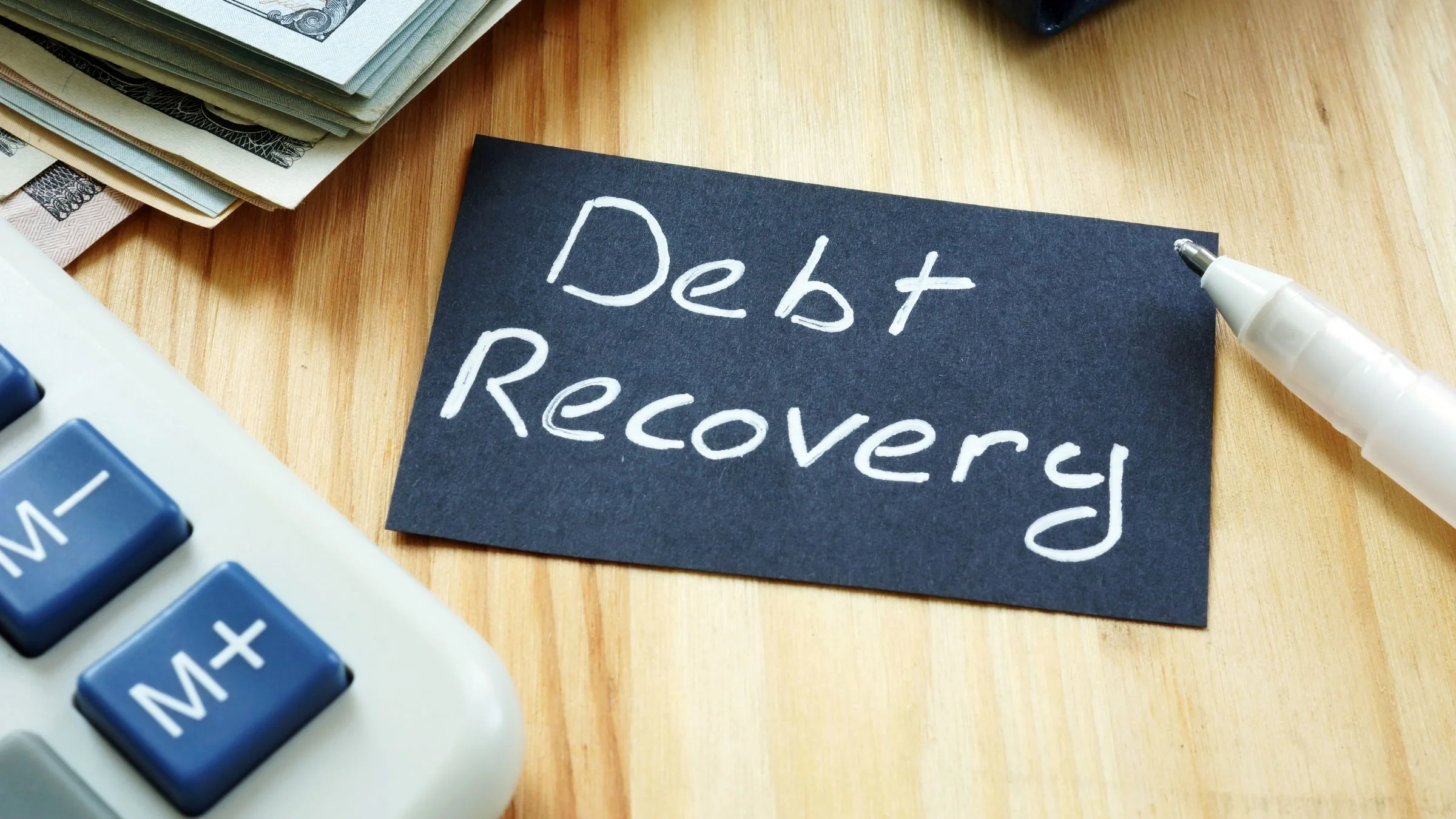 Card reading “Debt Recovery” on a desk beside cash, a calculator, a pen, and a wallet.