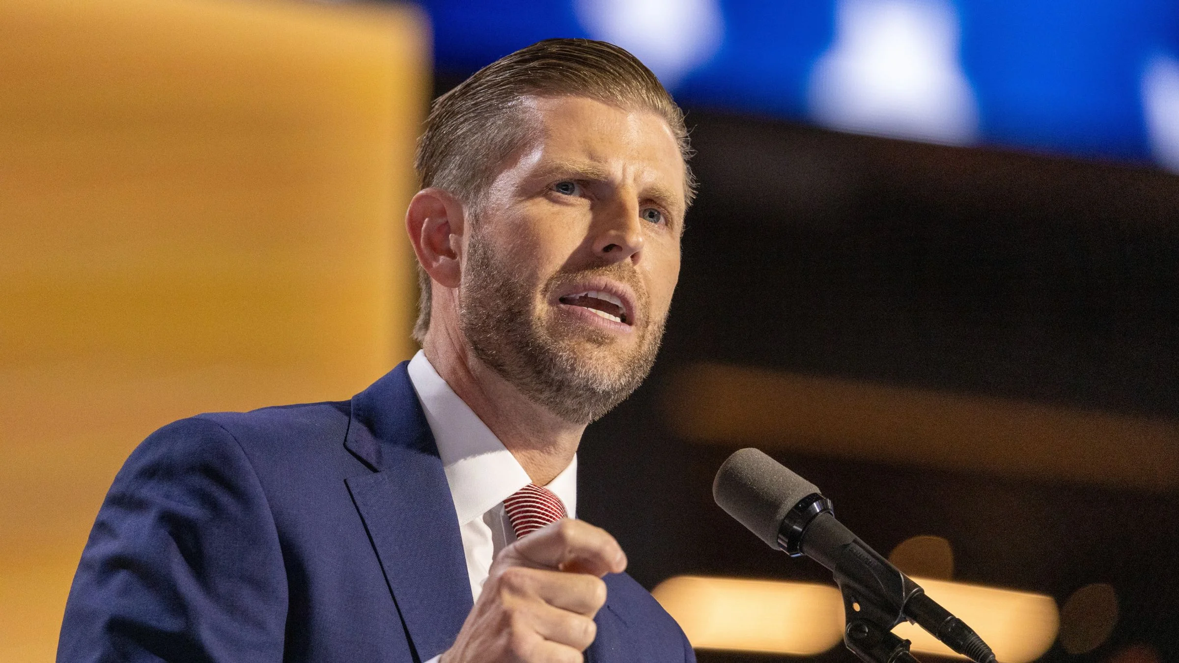Eric Trump speaking into a microphone at a podium while wearing a blue suit and red striped tie.