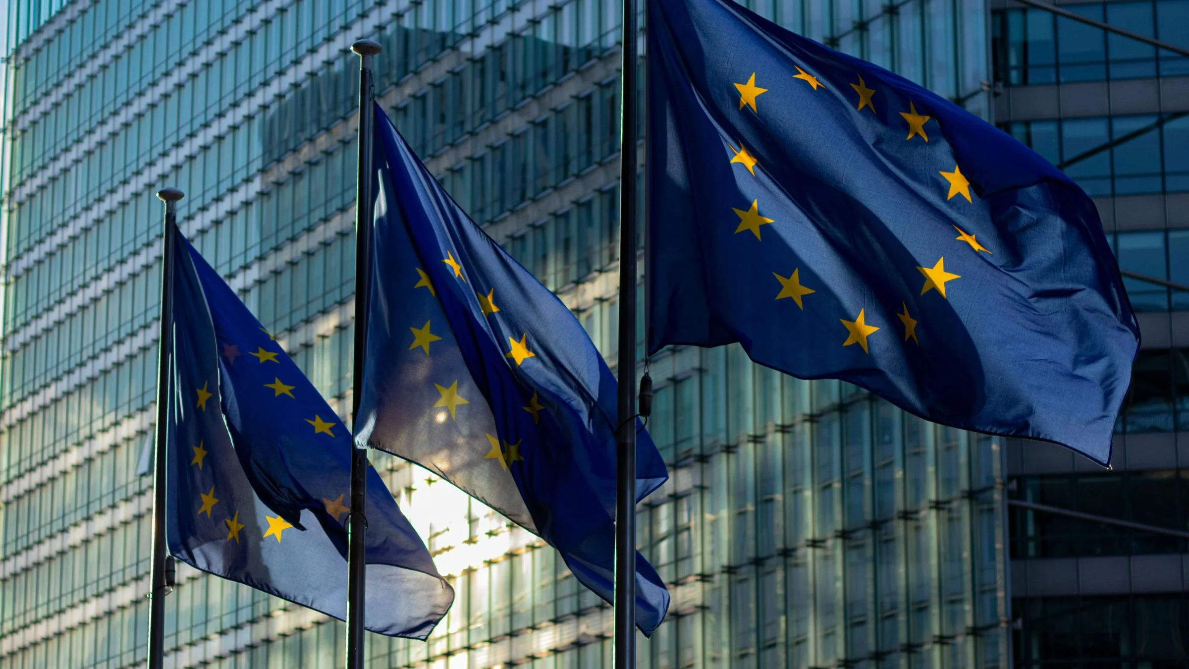 European Union flags flying outside a modern glass office building in an urban setting.