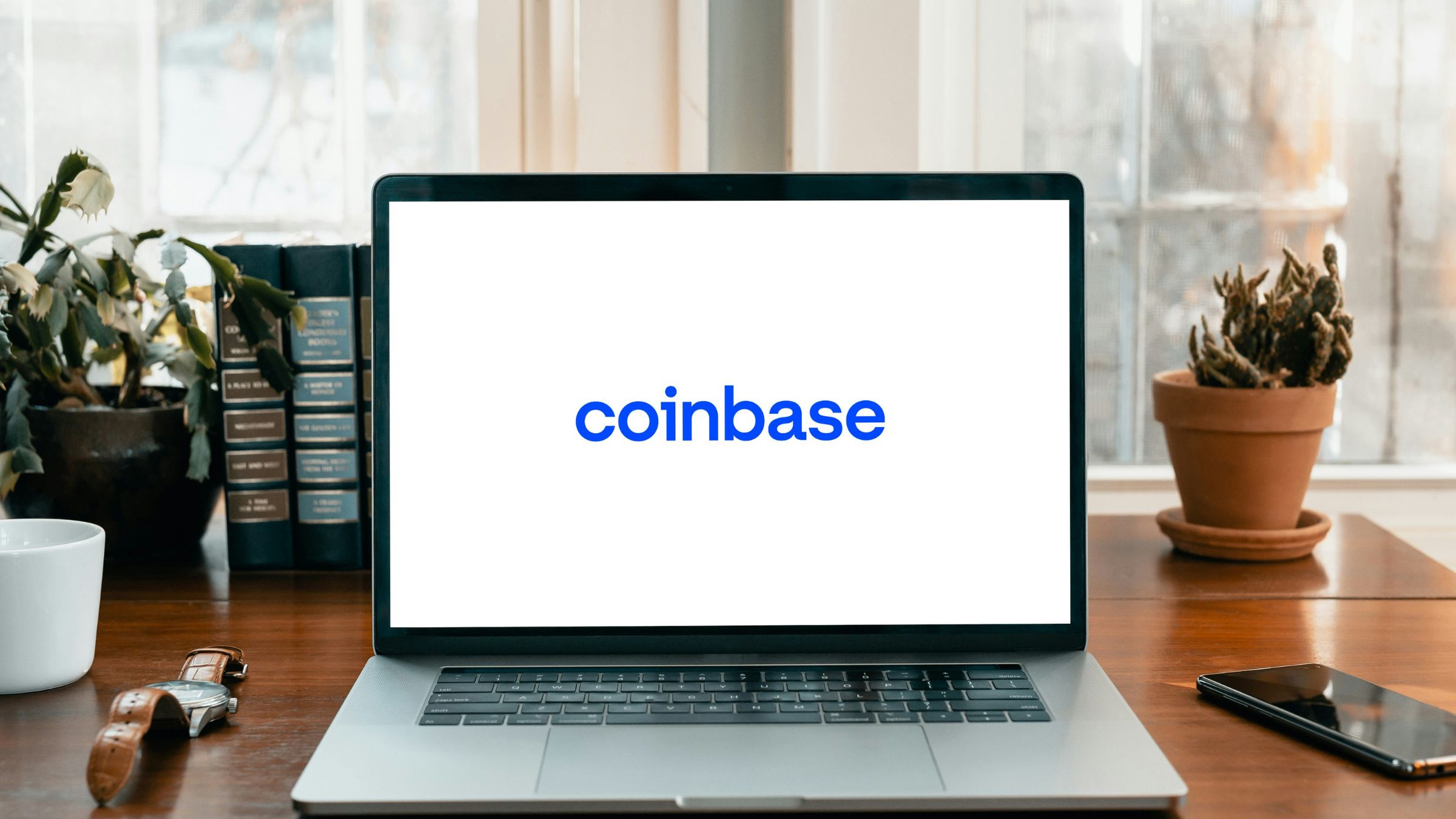 Laptop displaying the Coinbase logo on a desk with books, plants, and a smartphone nearby.