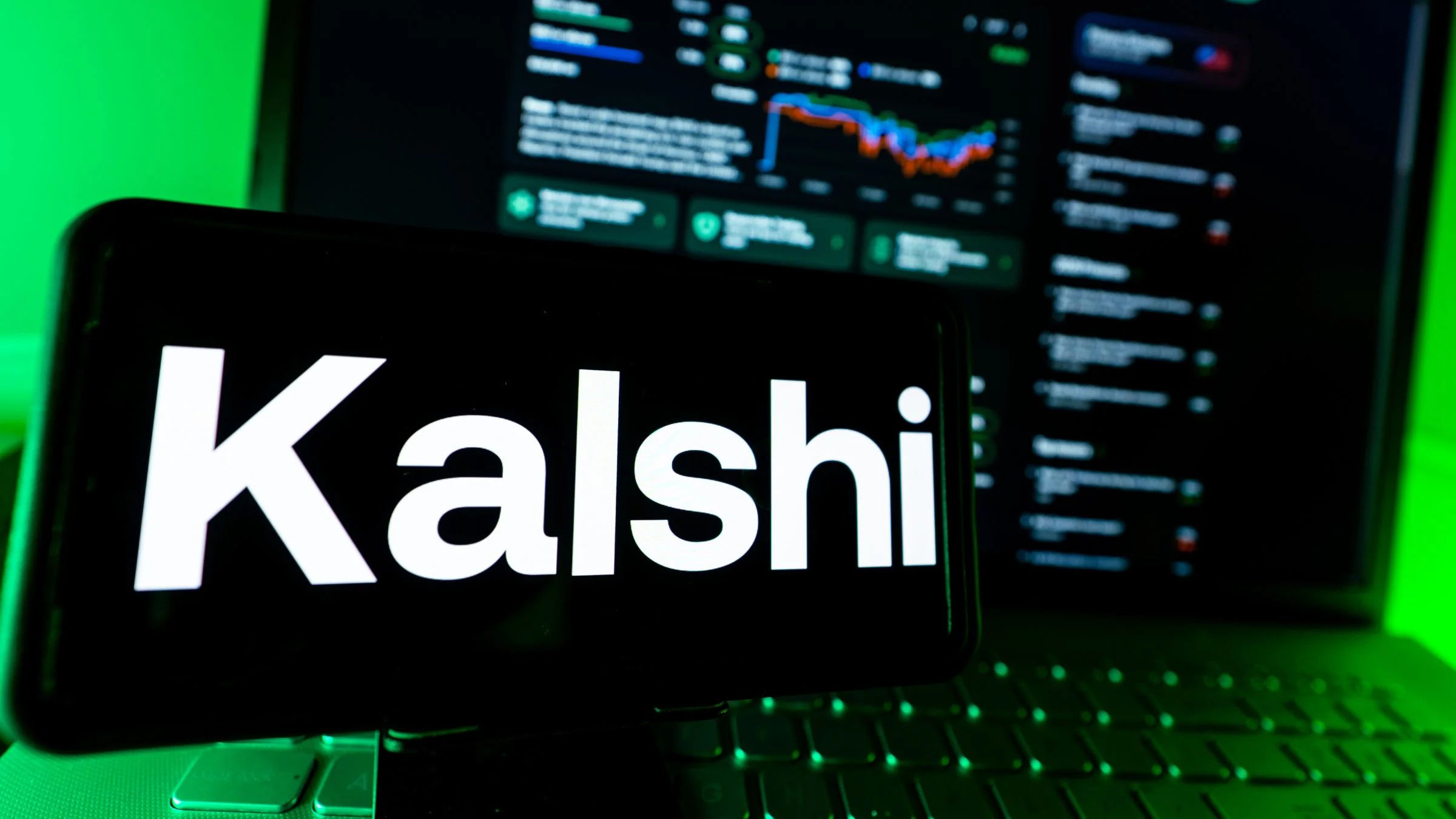 Smartphone displaying the Kalshi logo in front of a blurred laptop screen with market charts.