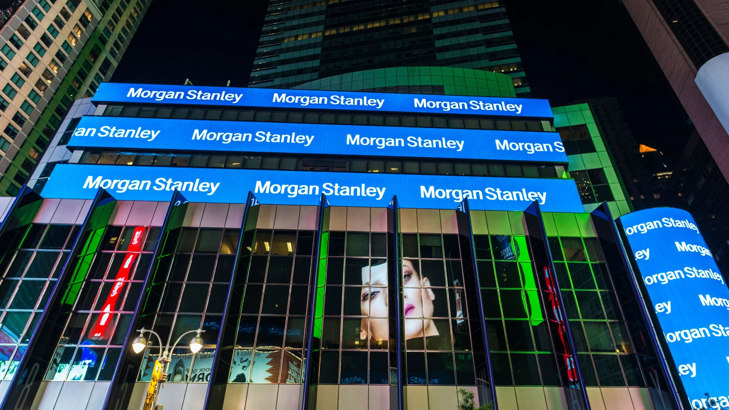 The illuminated facade of Morgan Stanley's headquarters in New York City at night, featuring multiple blue LED banners displaying the company's name across the glass building exterior.