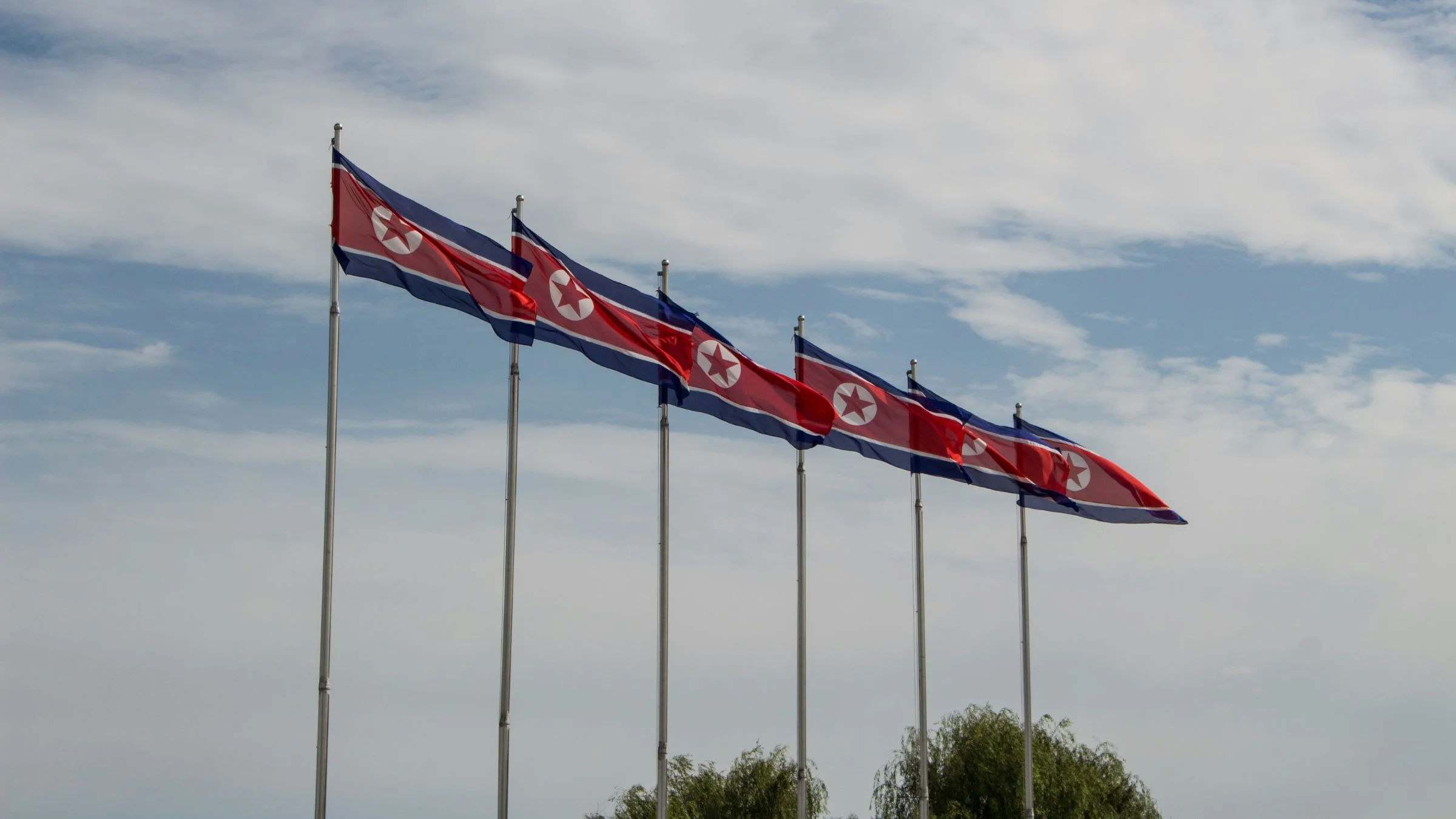 Several North Korean flags flying on tall flagpoles under a cloudy sky.