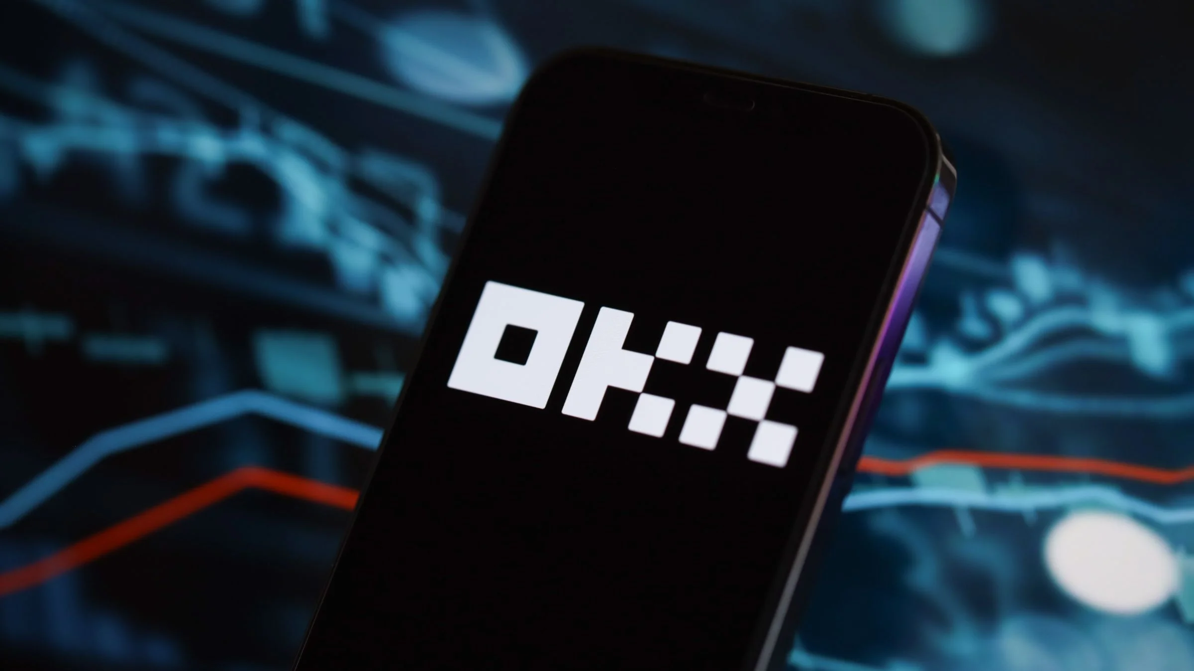 A smartphone displaying the OKX cryptocurrency exchange logo in white on a black screen, with blurred trading charts in the background.