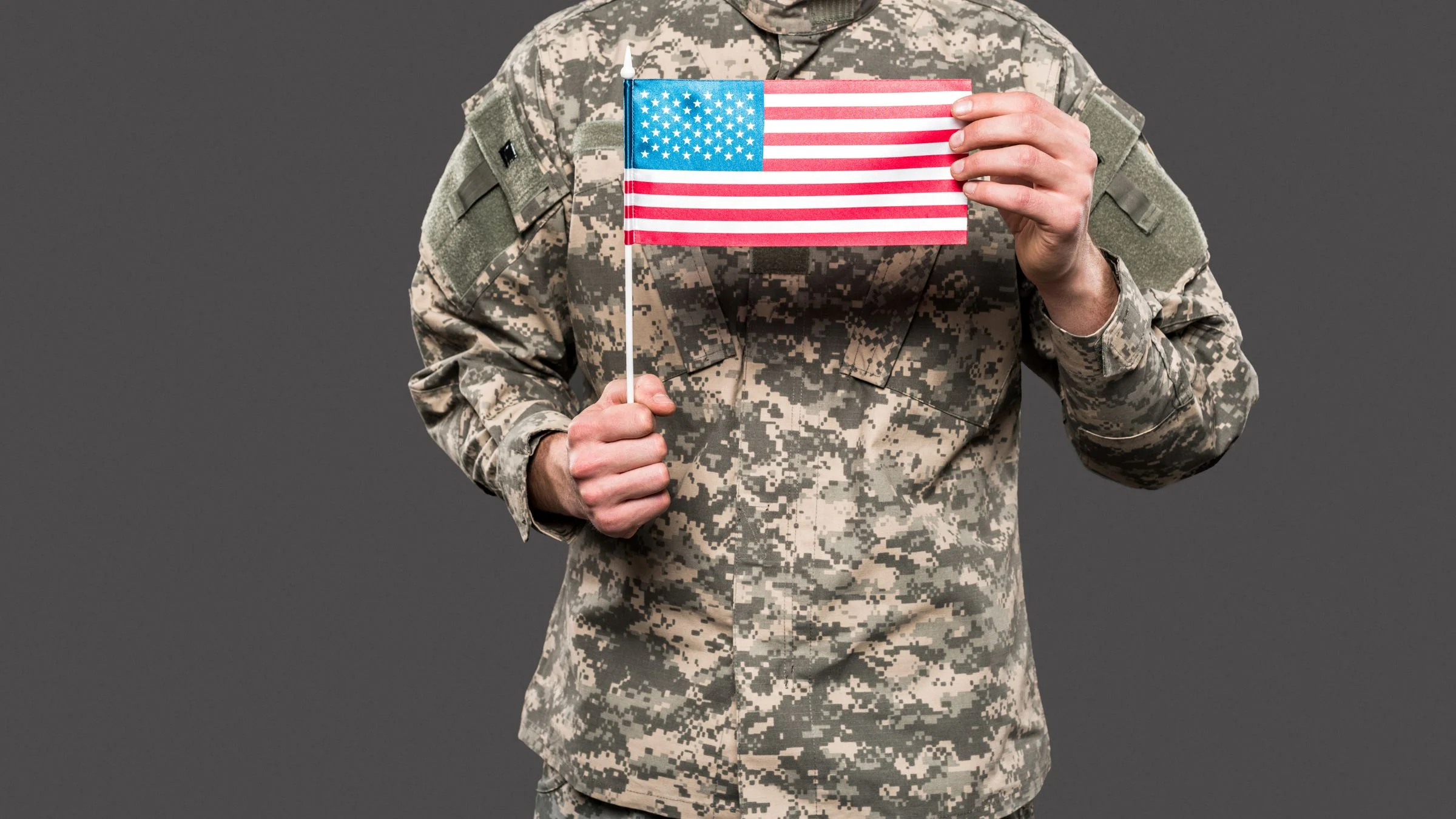 Person in camouflage uniform holding a small United States flag against a plain gray background.