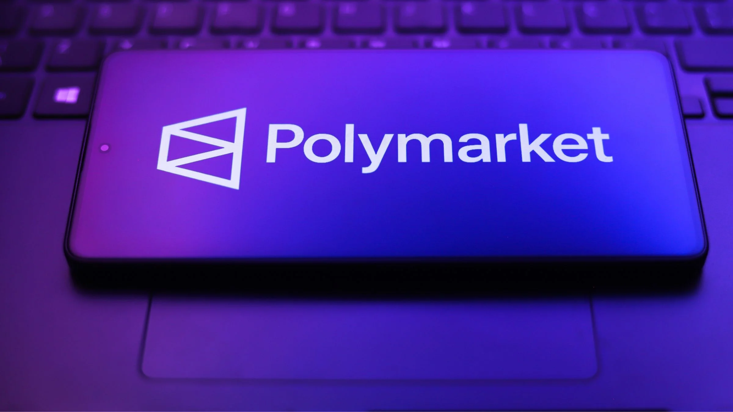 Smartphone displaying the Polymarket logo on a keyboard under purple and blue lighting.