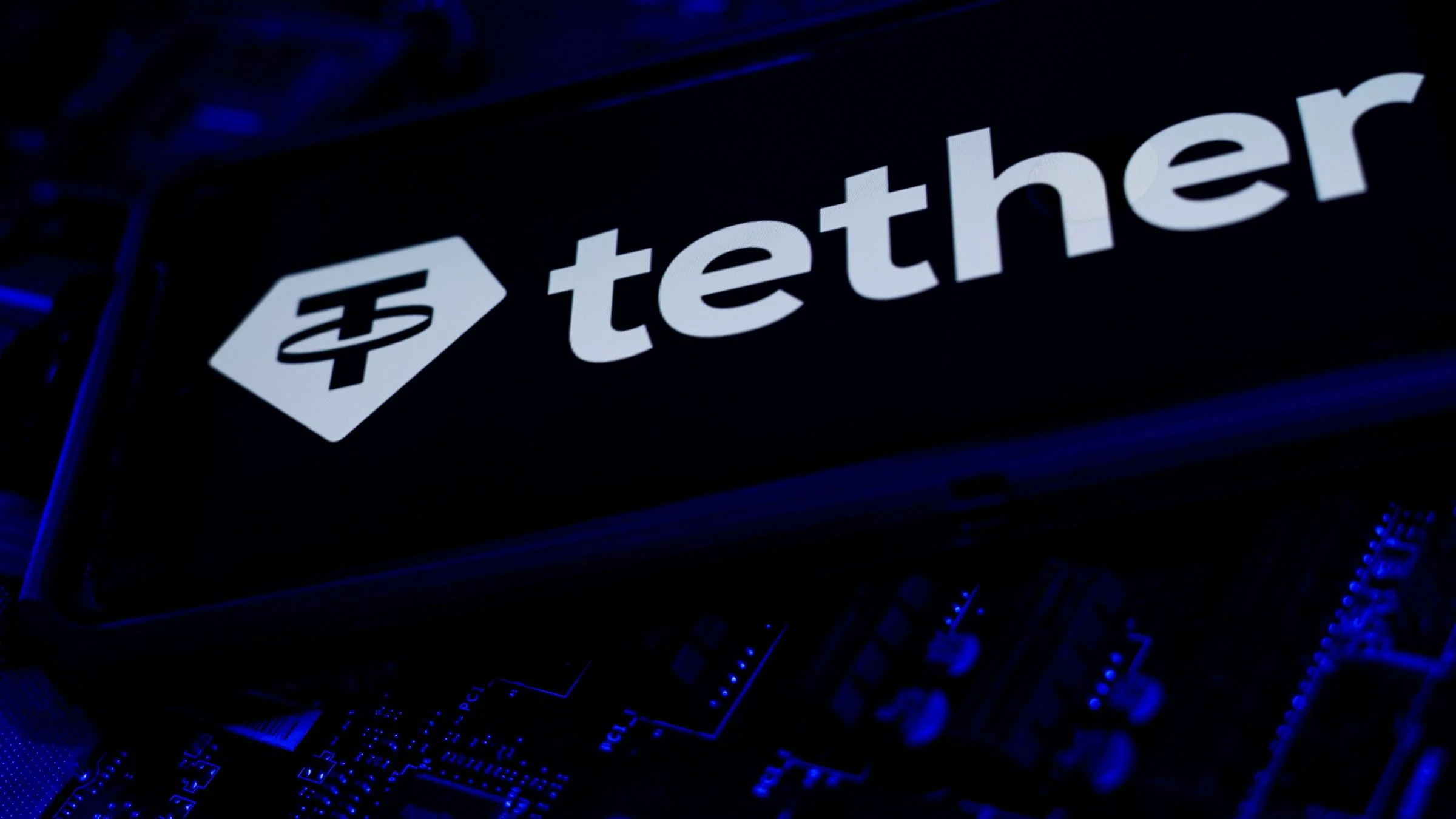 Tether logo displayed on a screen above electronic circuit components under blue lighting.