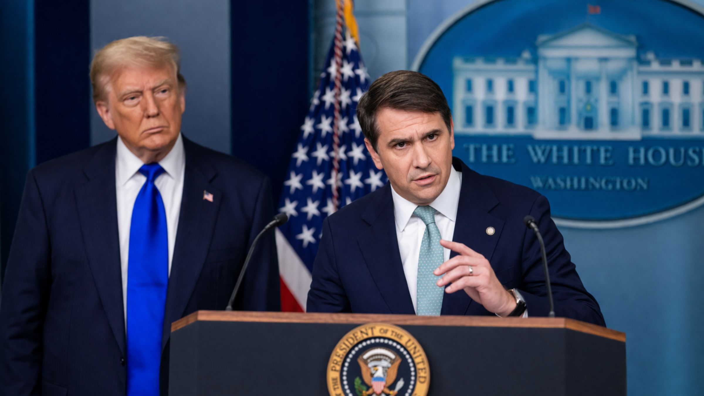 Todd Blanche speaks at a White House-style podium while Donald Trump stands behind him.