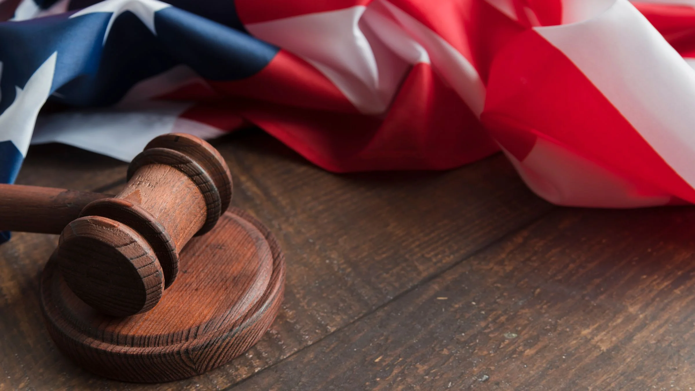 A wooden judge's gavel resting on a sound block on a rustic wooden table with a draped American flag in the background.