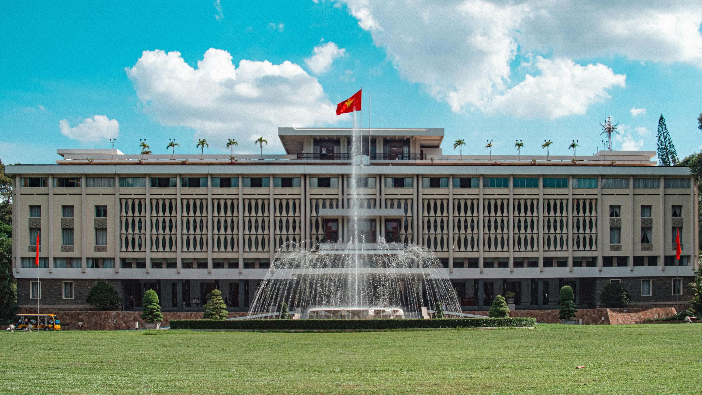 Large government-style building in Vietnam with a red national flag on top and a fountain in front under a bright blue sky.