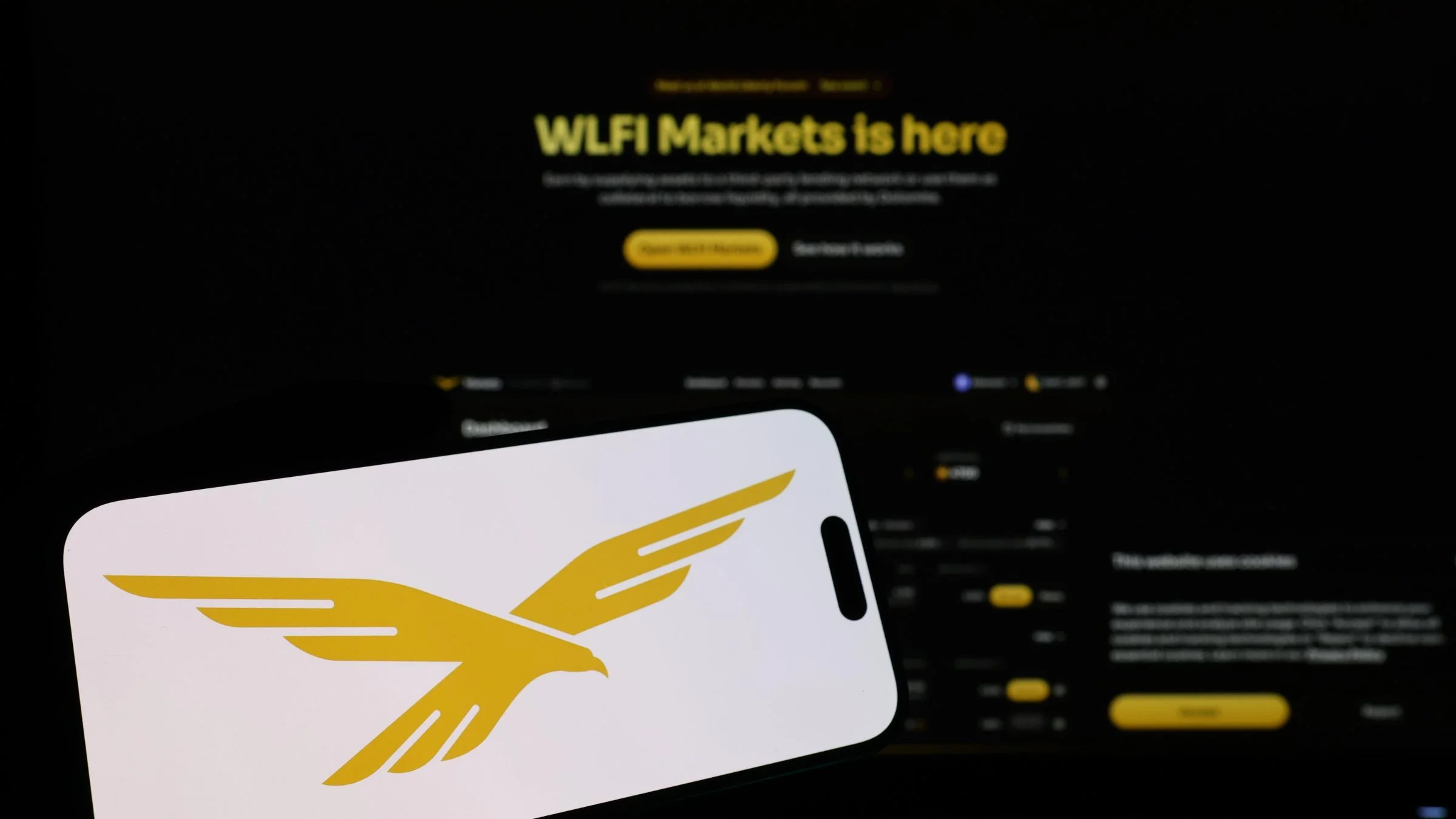 Smartphone displaying a gold eagle logo in front of a dark WLFI Markets interface on a computer screen.