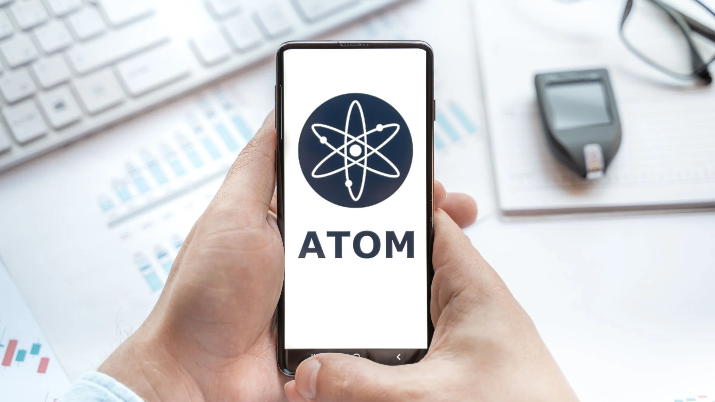 Hands holding a smartphone displaying the Cosmos ATOM cryptocurrency logo above a desk with financial charts and a keyboard