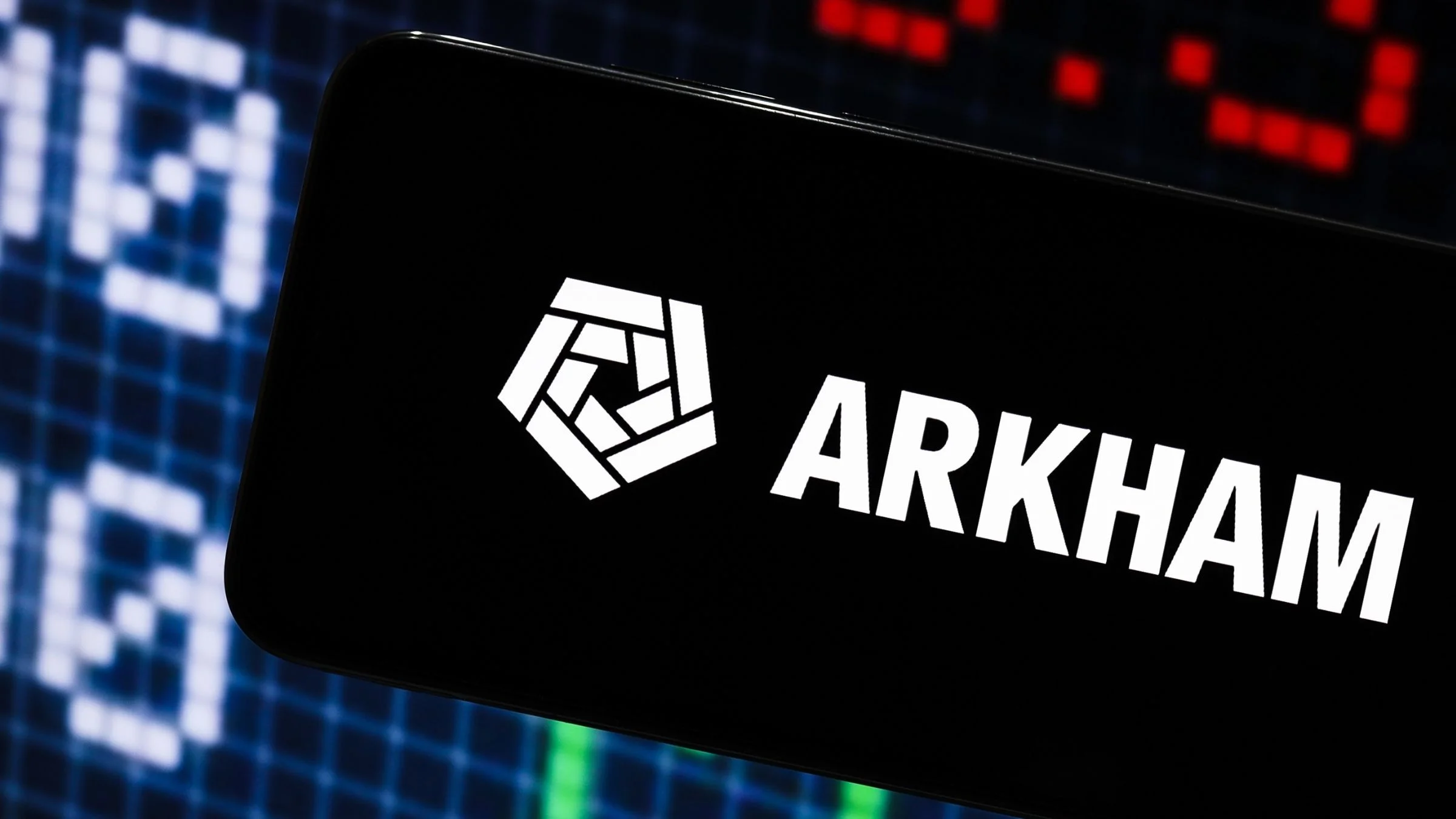 Arkham logo displayed on a smartphone screen with blurred red and green market data in the background.