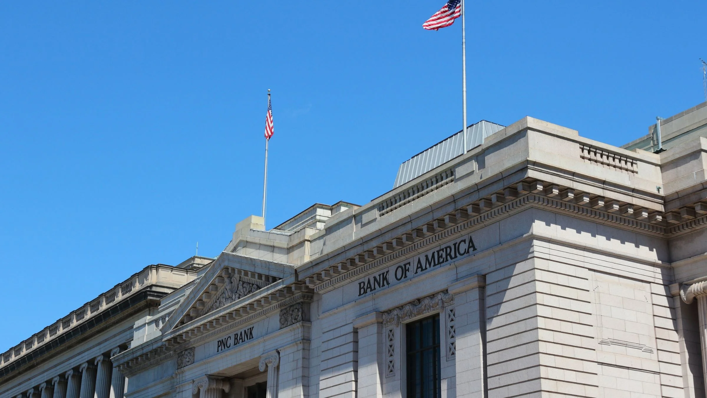 Exterior of a Bank of America building with U.S. flags flying above the facade under a clear sky.