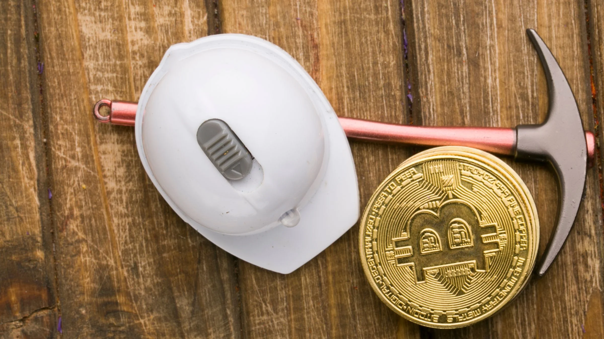 Gold Bitcoin coin beside a pickaxe and computer mouse on a wooden surface, symbolizing crypto mining.