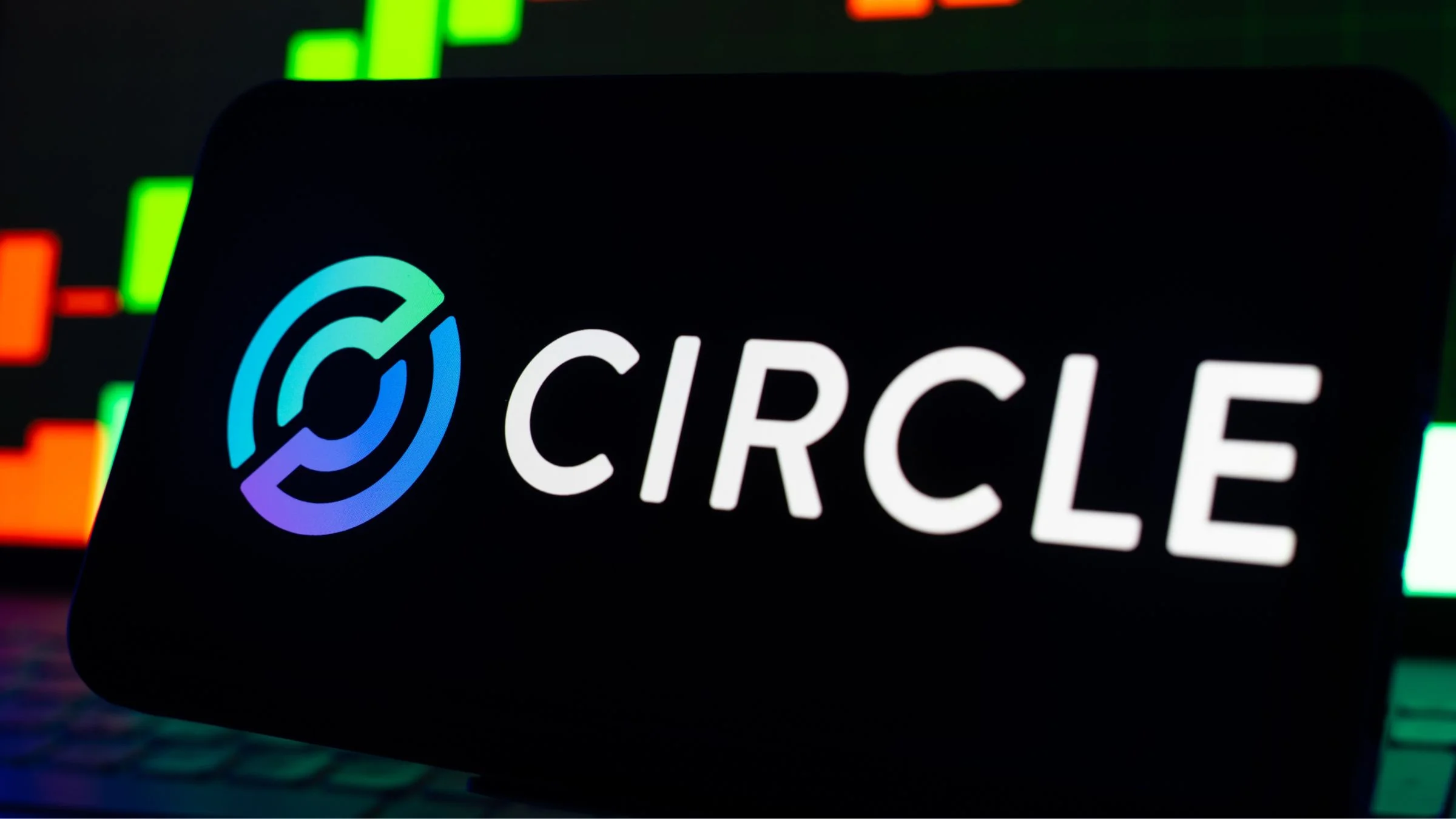 Circle logo displayed on a screen in front of a blurred green and red candlestick trading chart.