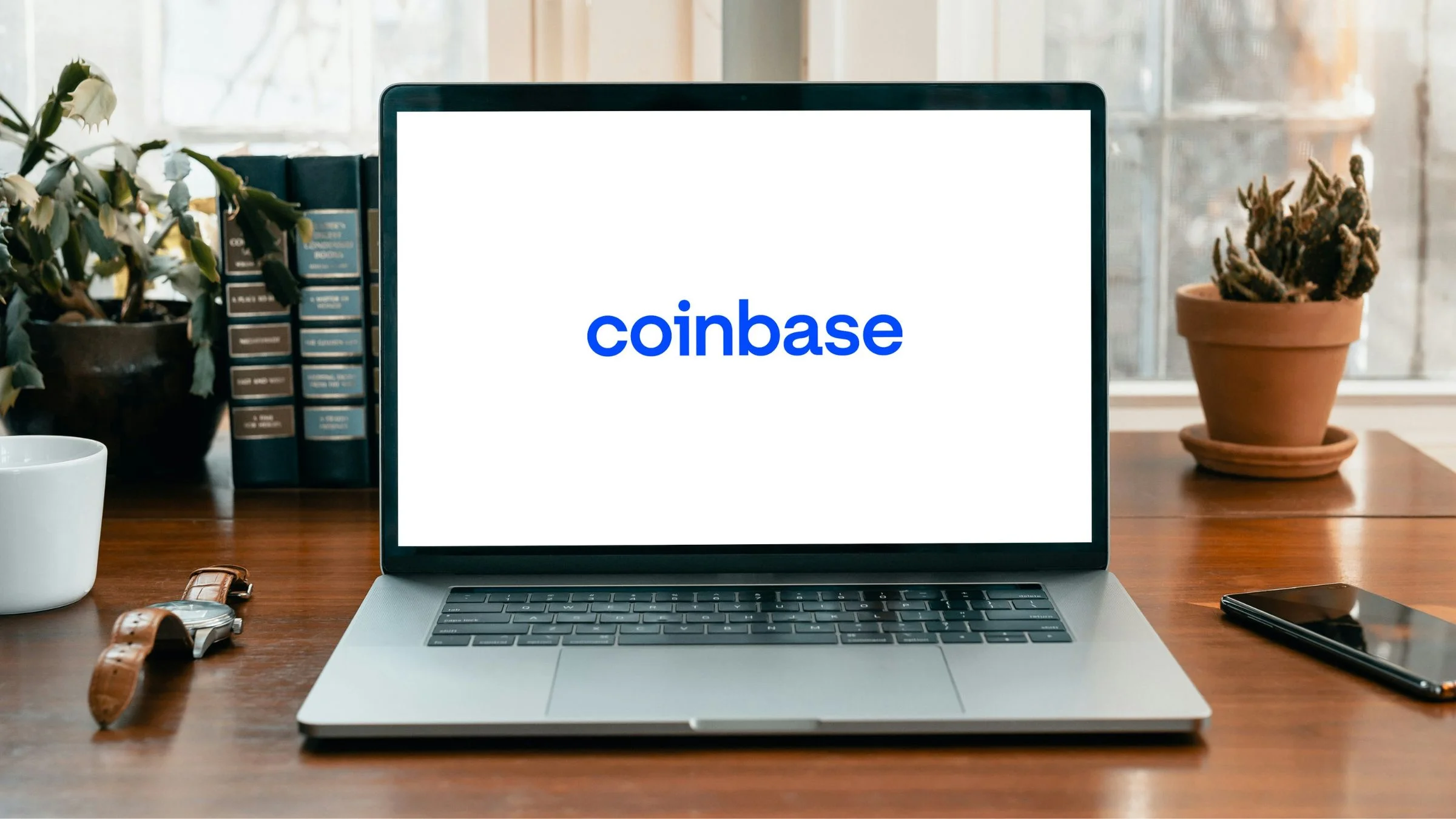 Laptop on a desk displaying the Coinbase logo, with books, plants, a mug, and a smartphone nearby.