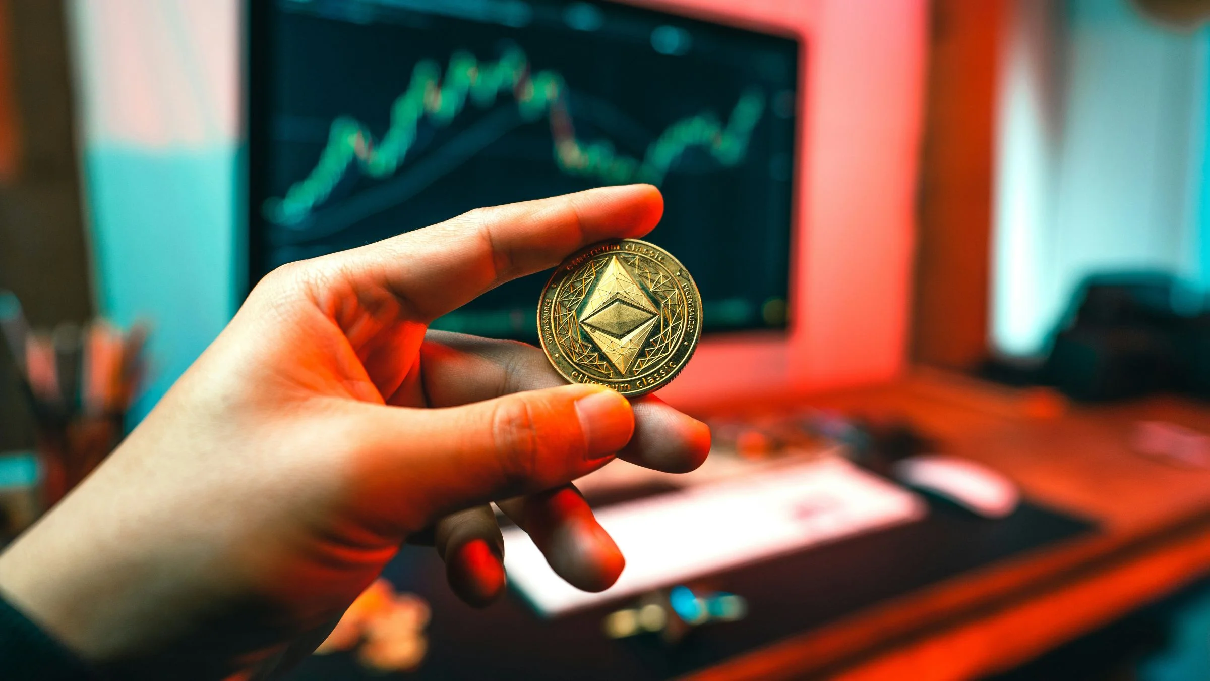 Hand holding an Ethereum Classic token in front of a blurred computer screen showing a market chart.