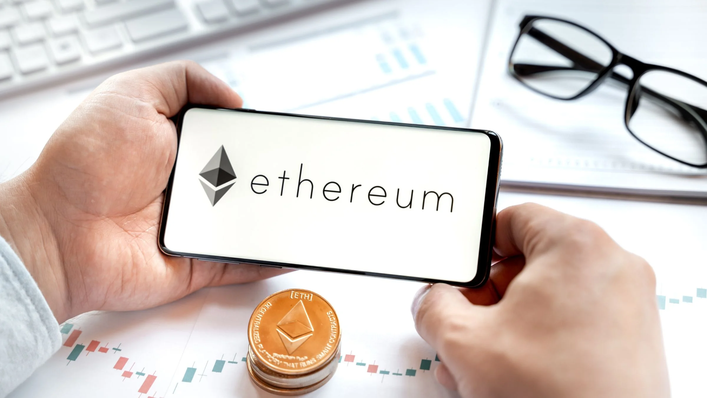 Ethereum logo shown on a smartphone screen above cryptocurrency market charts and an ETH token