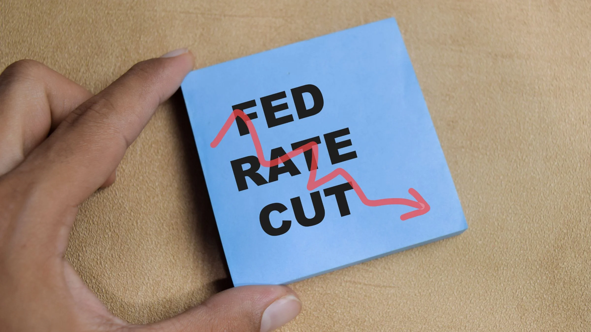 Hand holding a blue sticky note reading “FED RATE CUT” with a red downward arrow drawn across it.