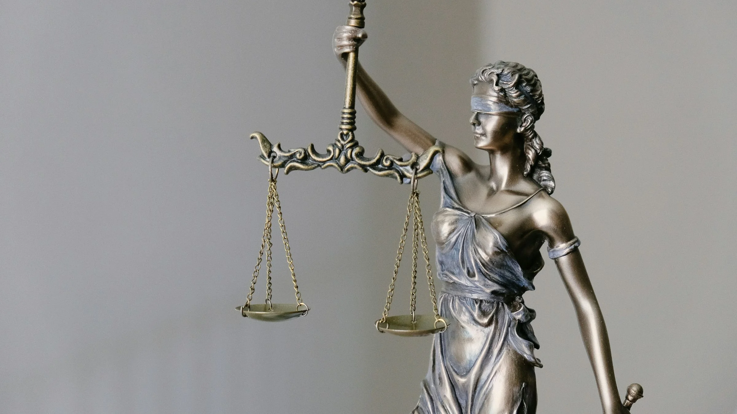 Statue of Lady Justice holding scales, with blindfold and sword, against a plain indoor background.