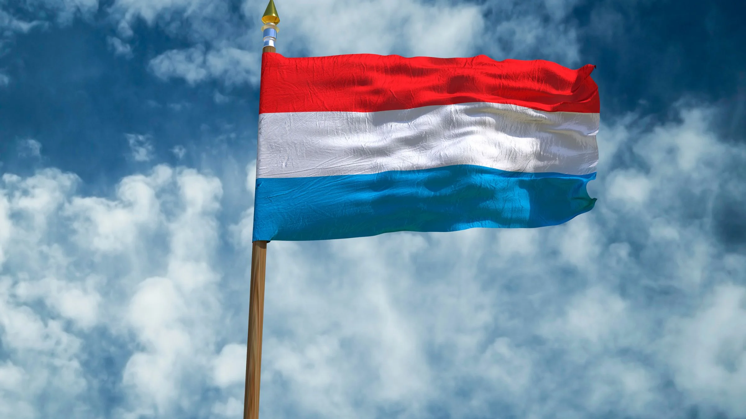 Luxembourg flag waving on a flagpole against a cloudy blue sky in bright daylight.