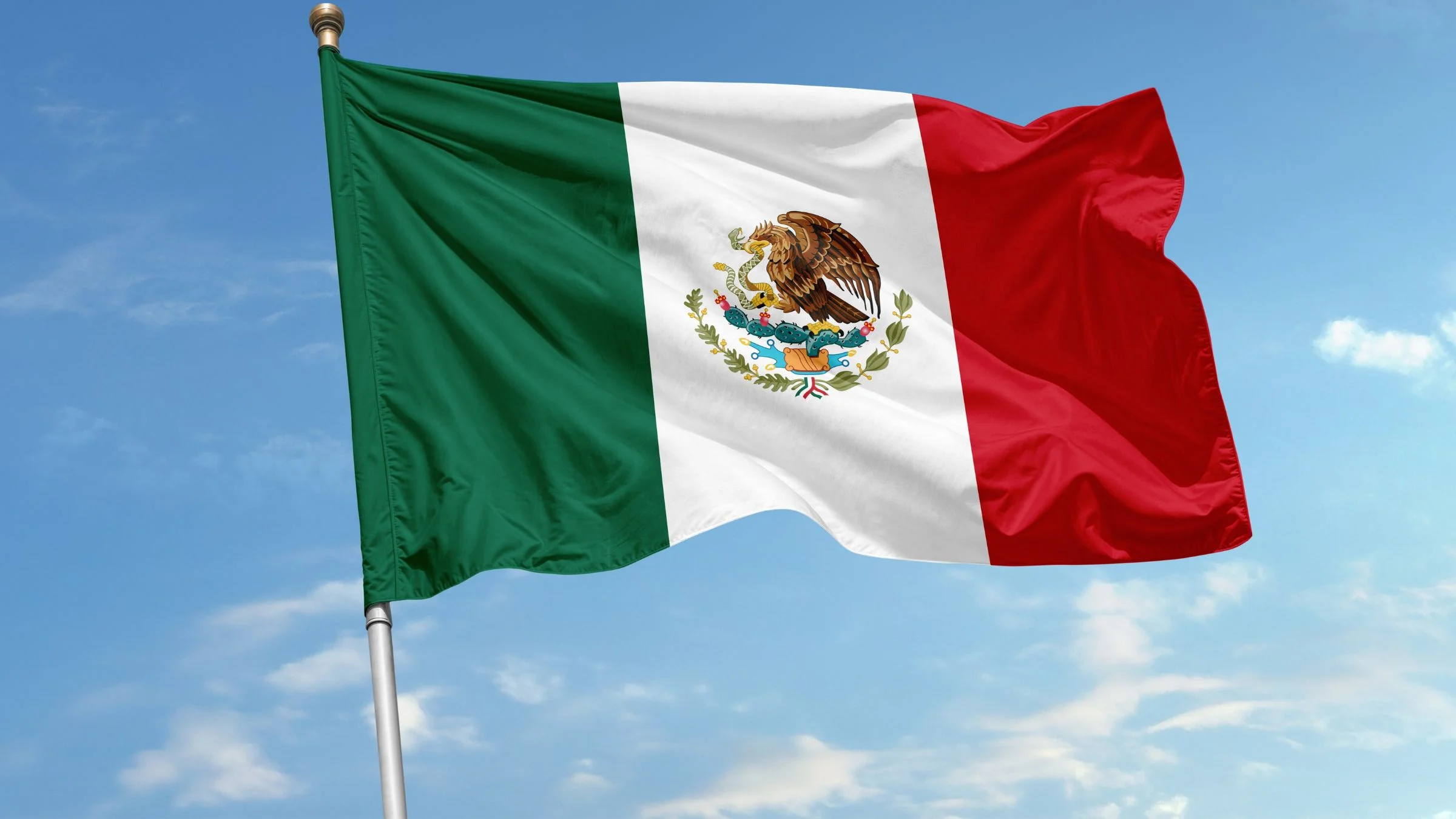 Mexican flag waving on a flagpole against a blue sky with light clouds.