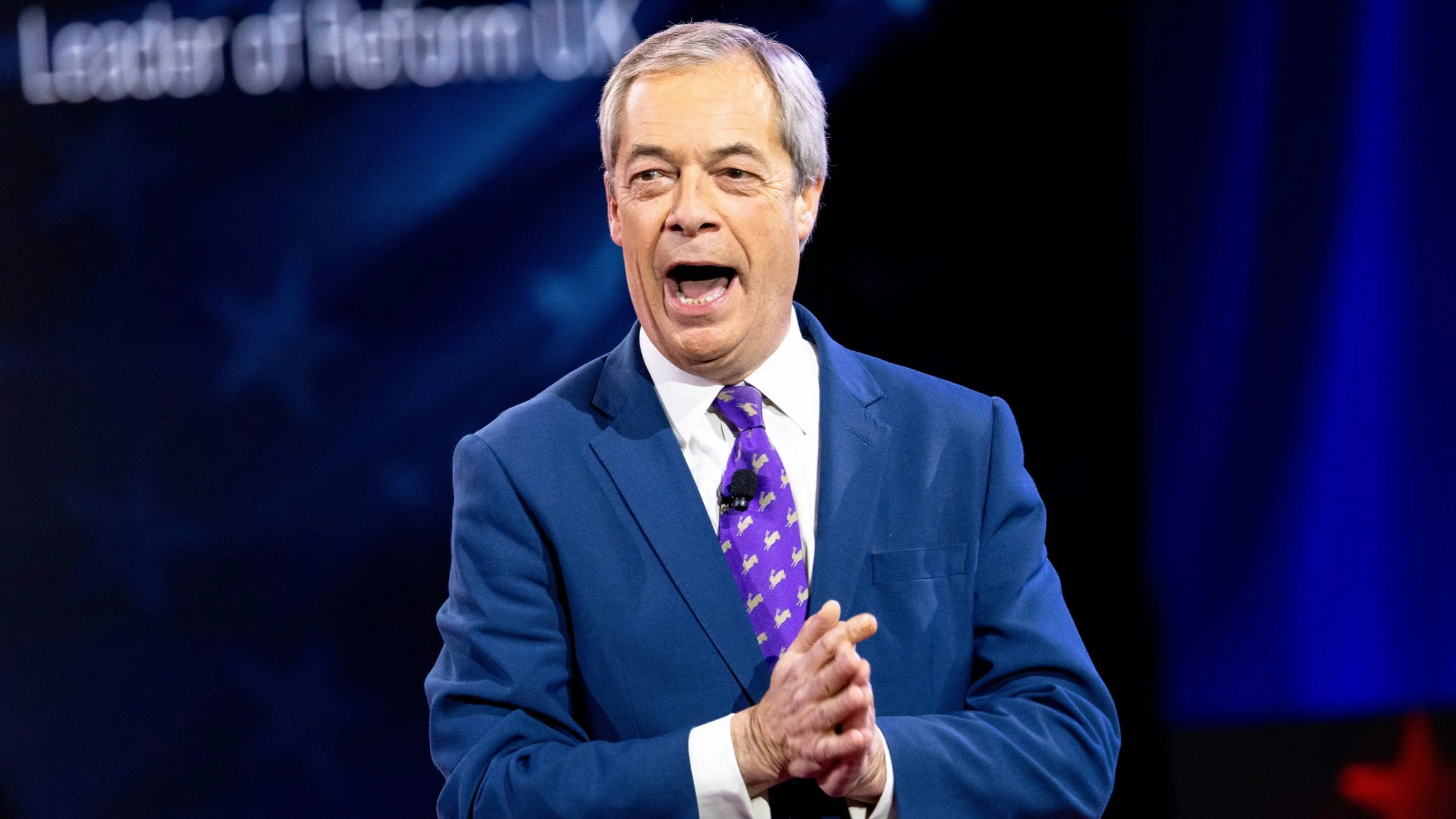 Man in a blue suit and purple tie speaking energetically on a dark stage with text reading "Leader of Reform UK" visible in the upper left