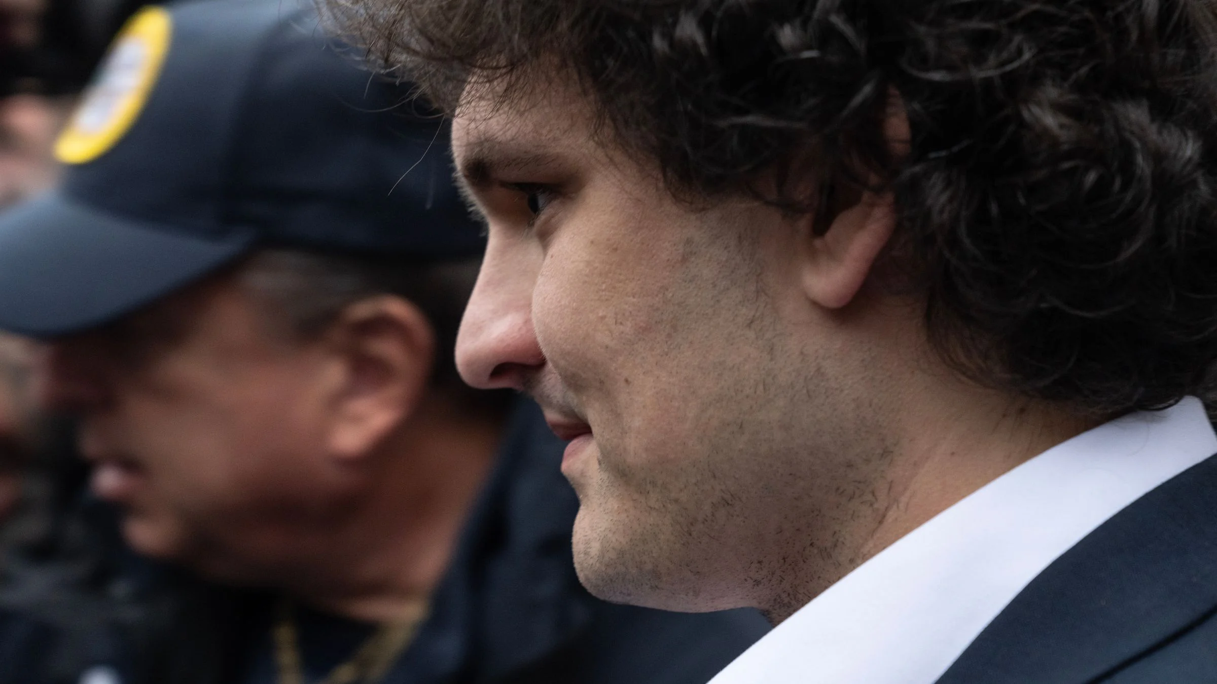 Close-up side profile of a Sam Bankman-Fried in a suit standing beside a police officer in a crowd.