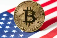 A gold physical Bitcoin coin with a large "B" symbol resting upright on a folded American flag.