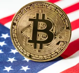 A gold physical Bitcoin coin with a large "B" symbol resting upright on a folded American flag.