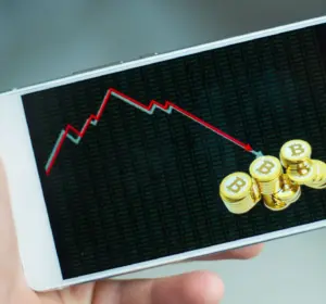 Hand holding a smartphone showing a falling chart arrow and Bitcoin coin graphics on screen.