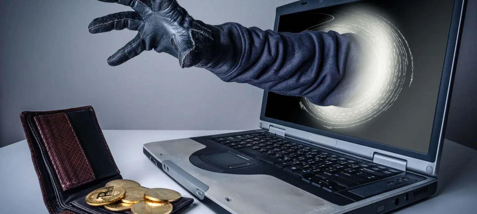 Gloved hand reaching from a laptop toward an open wallet holding Bitcoin coins.