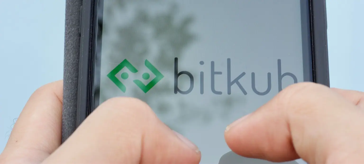 Hands holding a mobile device displaying the Bitkub logo against a softly blurred outdoor background.